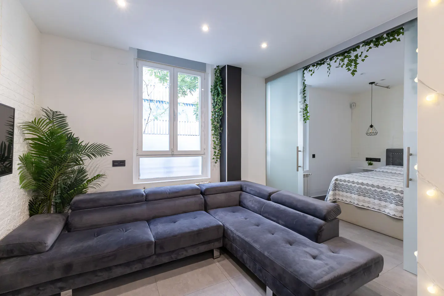 A modern apartment interior with a gray sectional sofa, a window, and a glass sliding door to the bedroom.