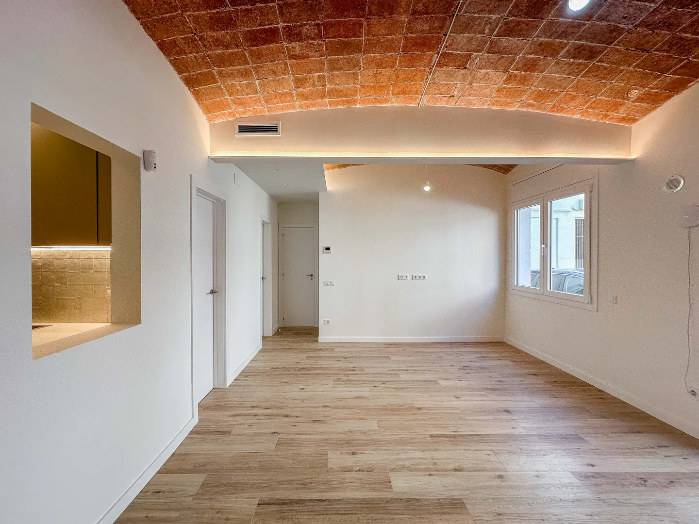 Bright, empty room with light wood floors, white walls, and a red brick vaulted ceiling. A window lets in natural light.