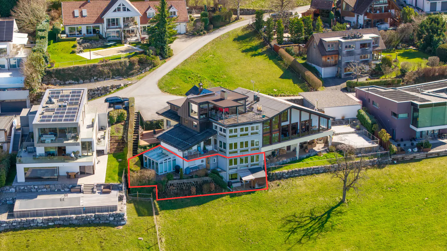 Aerial view of a multi-story house with a red outline, set on a green hillside with other houses visible in the background.