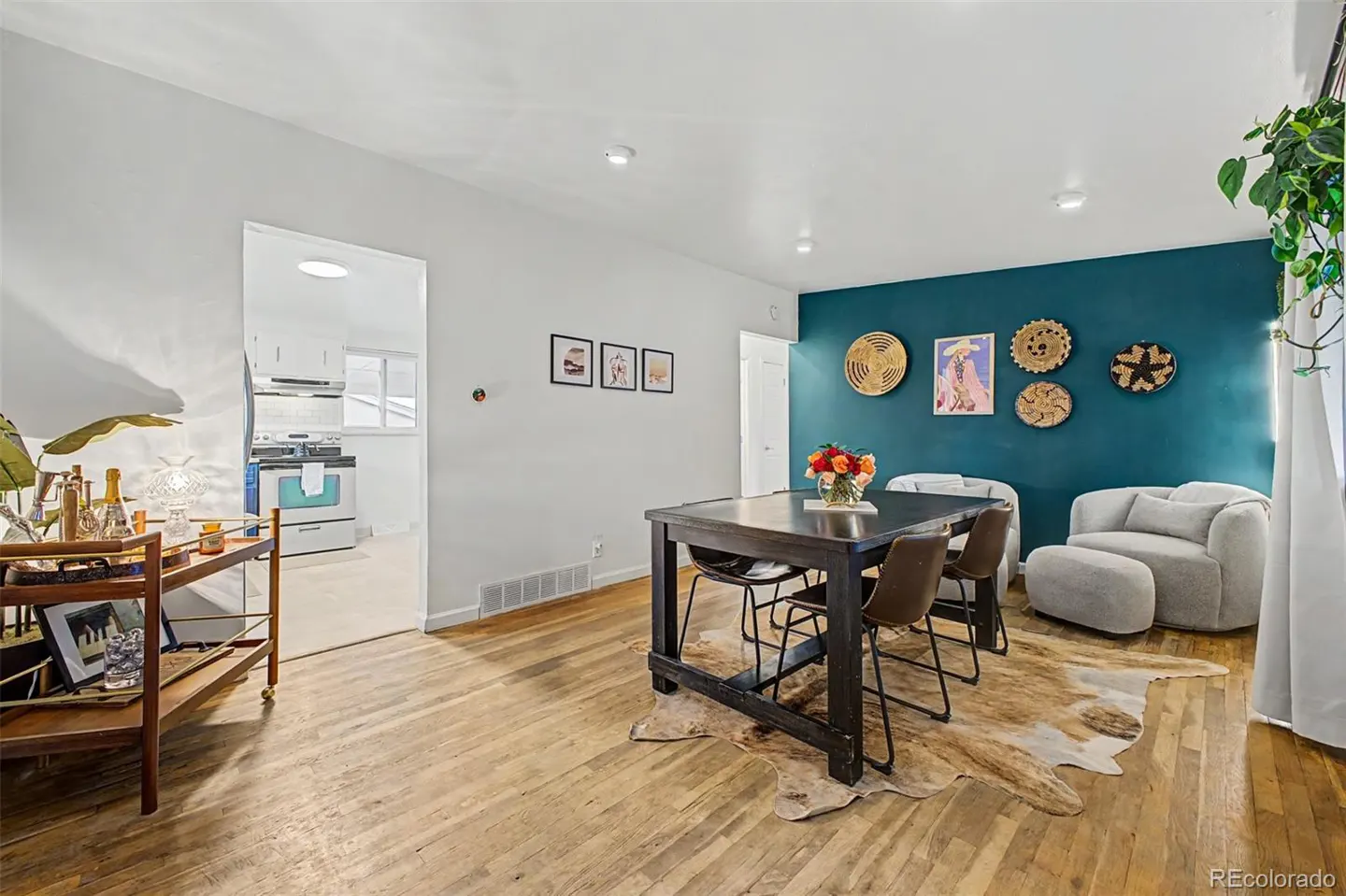 Dining room with dark table, chairs, and cowhide rug against a teal accent wall, with a bar cart and kitchen view.