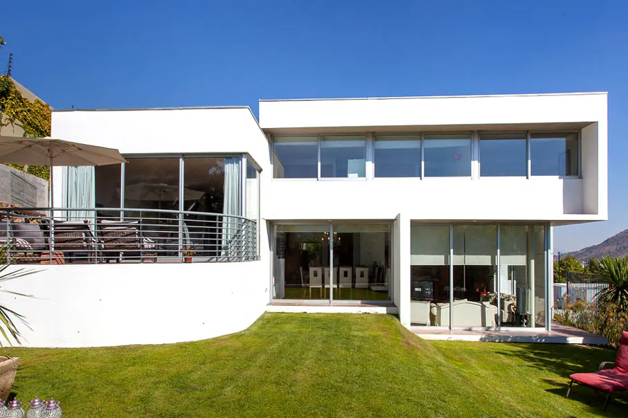 Modern white house with large windows and a curved balcony overlooking a green lawn under a clear blue sky.