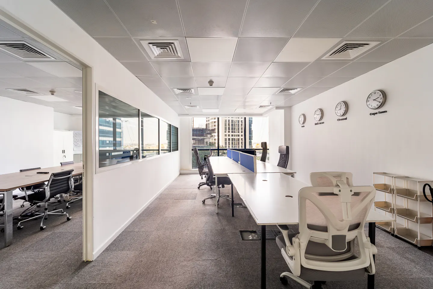 Bright office space with white walls, gray carpet, and white desks. Clocks showing different time zones hang on the wall. A conference room is visible through a doorway.