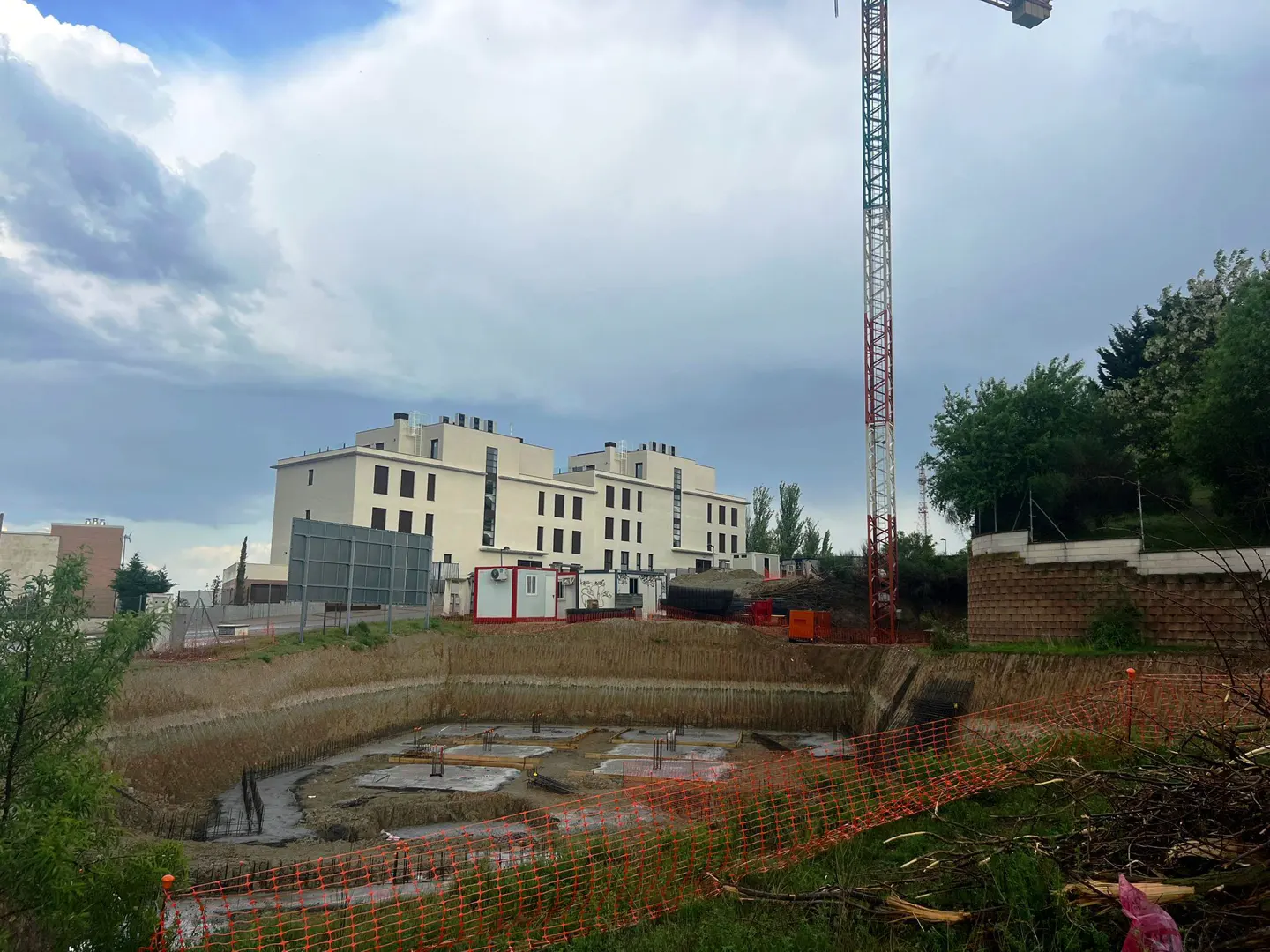 Construction site with a large pit, concrete foundations, and a tall crane against a cloudy sky. A white building is in the background.