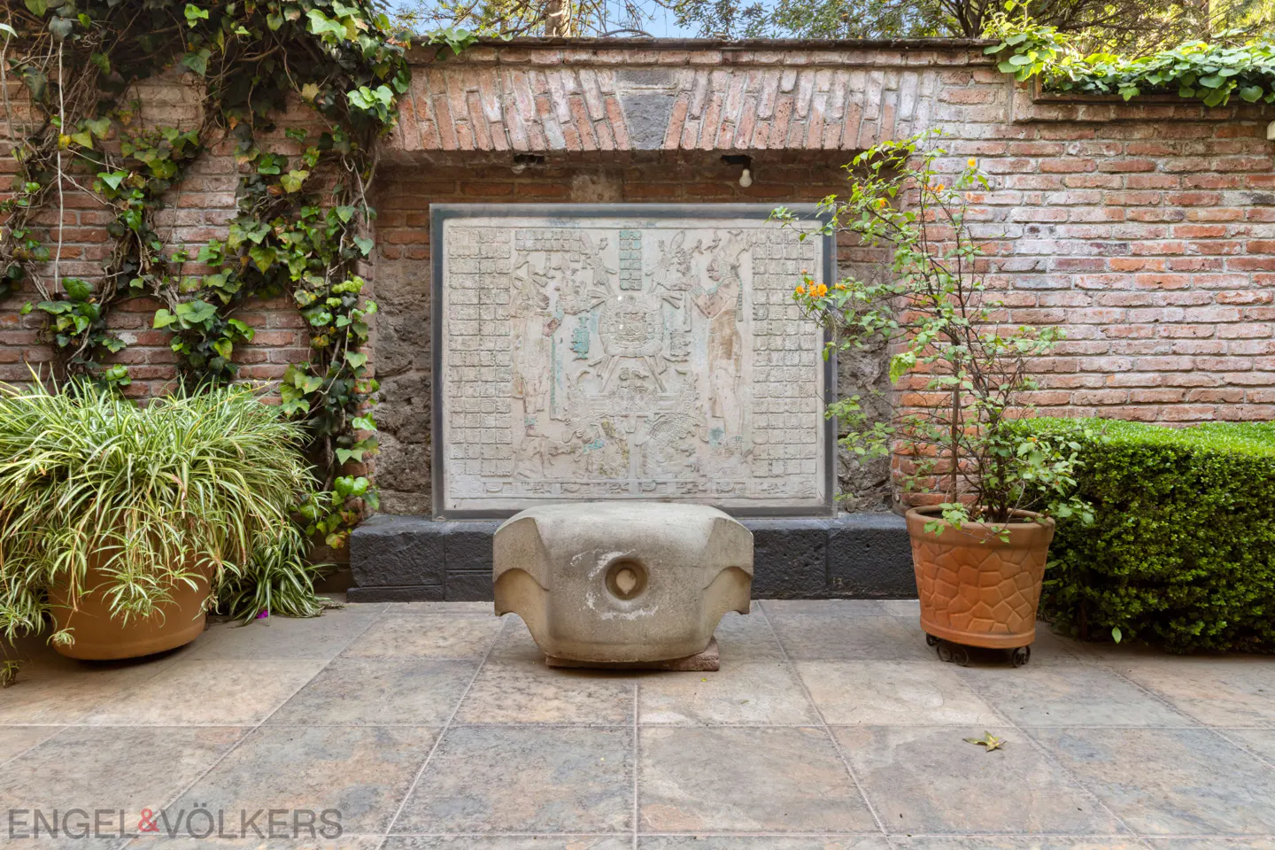 Outdoor patio with brick walls, ivy, stone bench, and a framed stone carving. Potted plants add greenery.