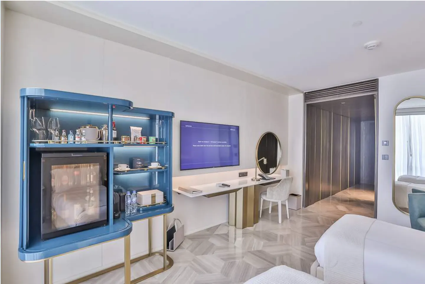 Hotel room interior with a blue mini-bar, TV, desk, chair, and a glimpse of the bed. The room has a modern design with white walls and herringbone floors.
