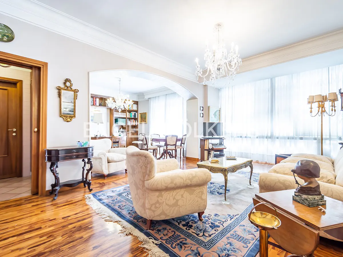 A bright living room with hardwood floors, a blue rug, and cream-colored furniture. A chandelier hangs from the ceiling.