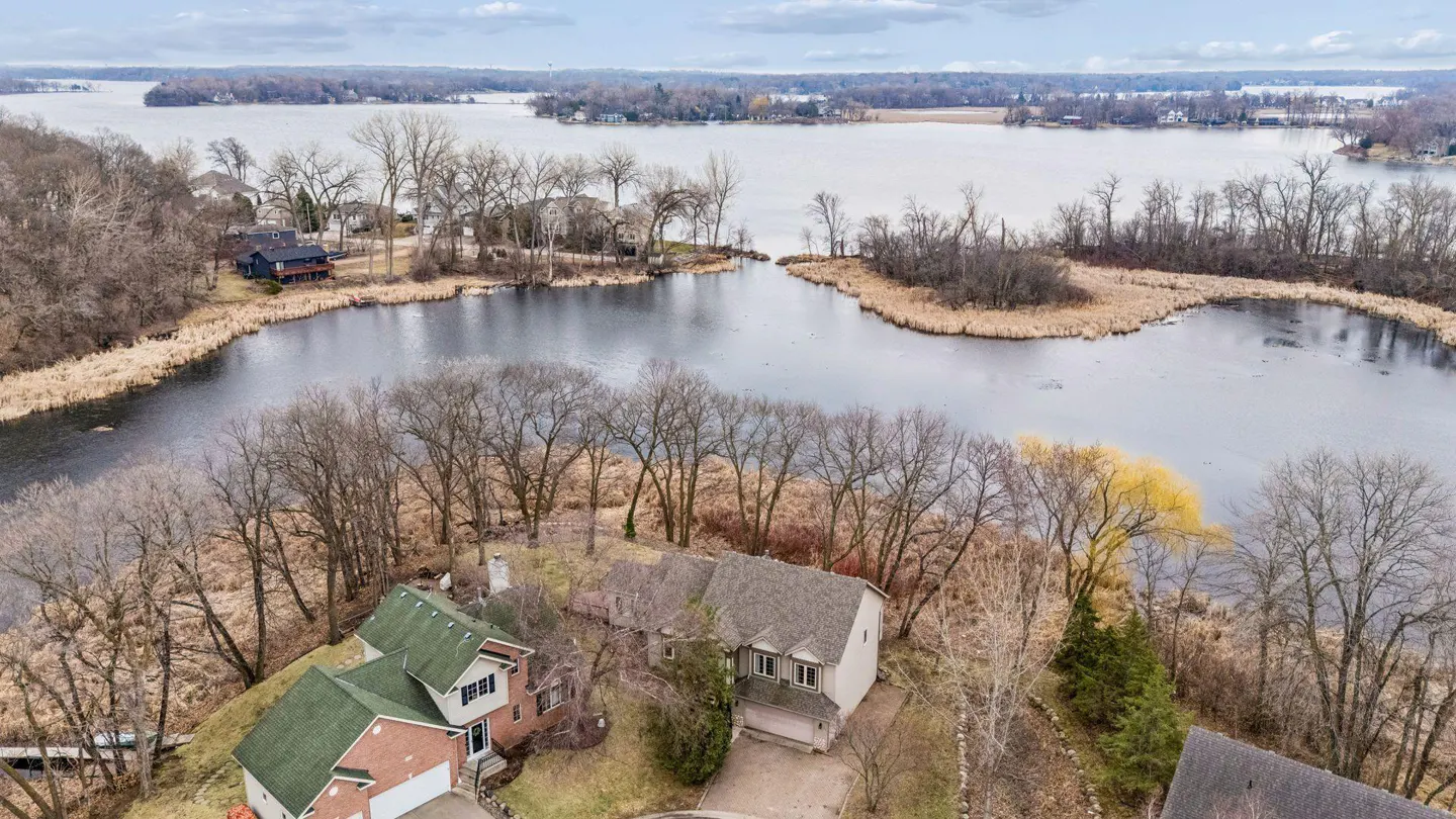 Aerial view of two houses near a lake with bare trees and a cloudy sky. One house has a green roof and the other has a gray roof.