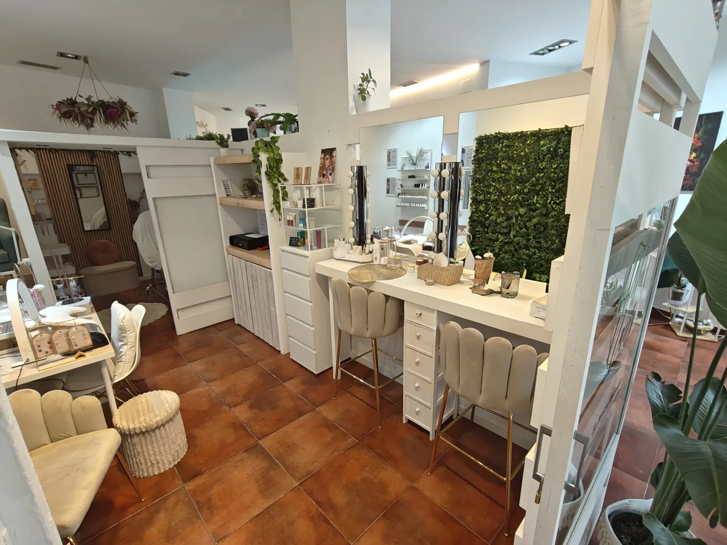 Interior of a beauty salon with white makeup stations, beige chairs, and mirrors with lights. Green plant wall in the background.