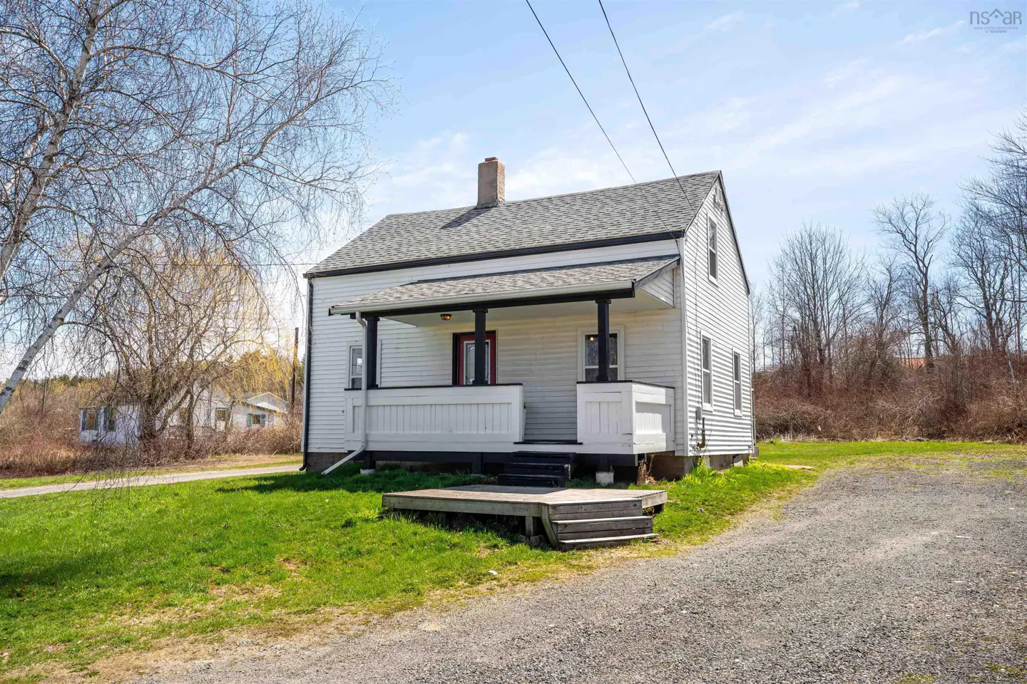 A white, two-story house with a gray roof and a small porch on a sunny day. A gravel driveway leads to the house.