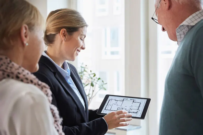 A real estate agent shows a floor plan on a tablet to an elderly couple.