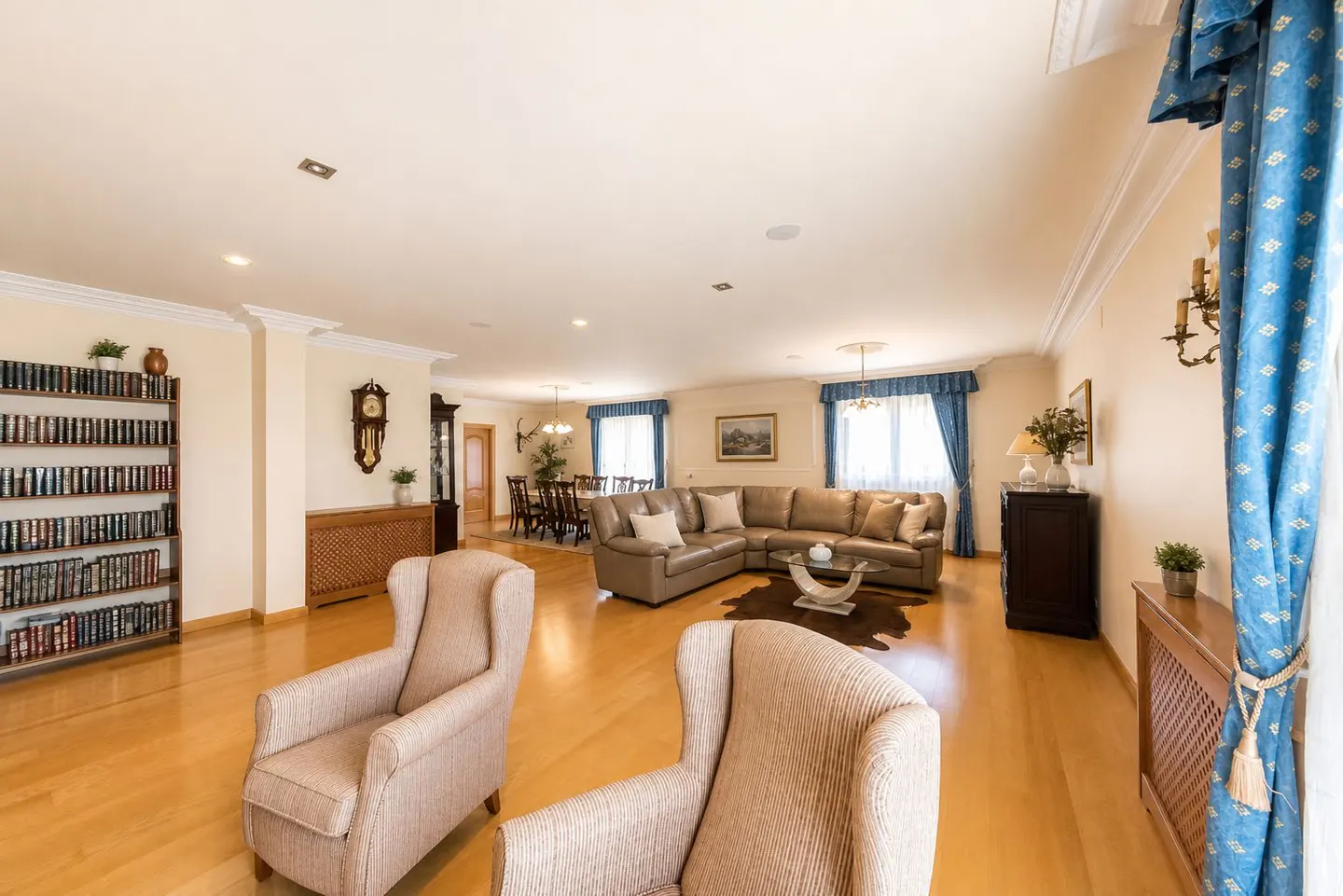 A spacious living room with hardwood floors, beige walls, and blue curtains. Two armchairs face a brown leather sectional sofa. A bookshelf and dining table are visible.