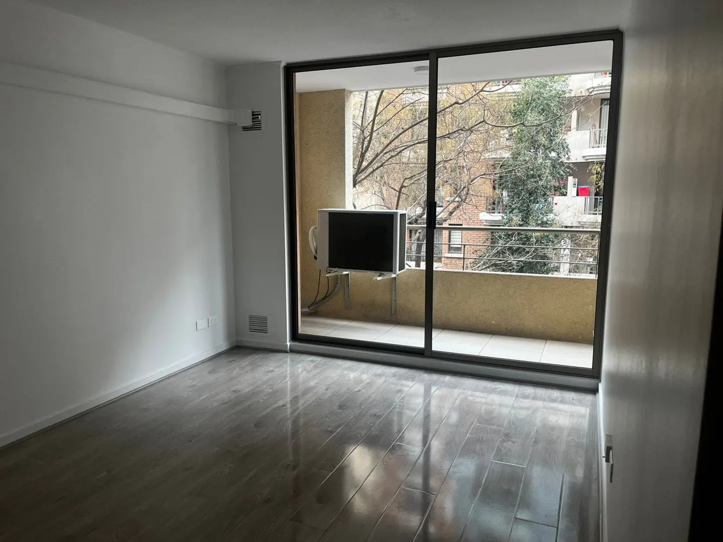 Empty room with gray wood floors, white walls, and a sliding glass door to a balcony with a view of trees and a building.