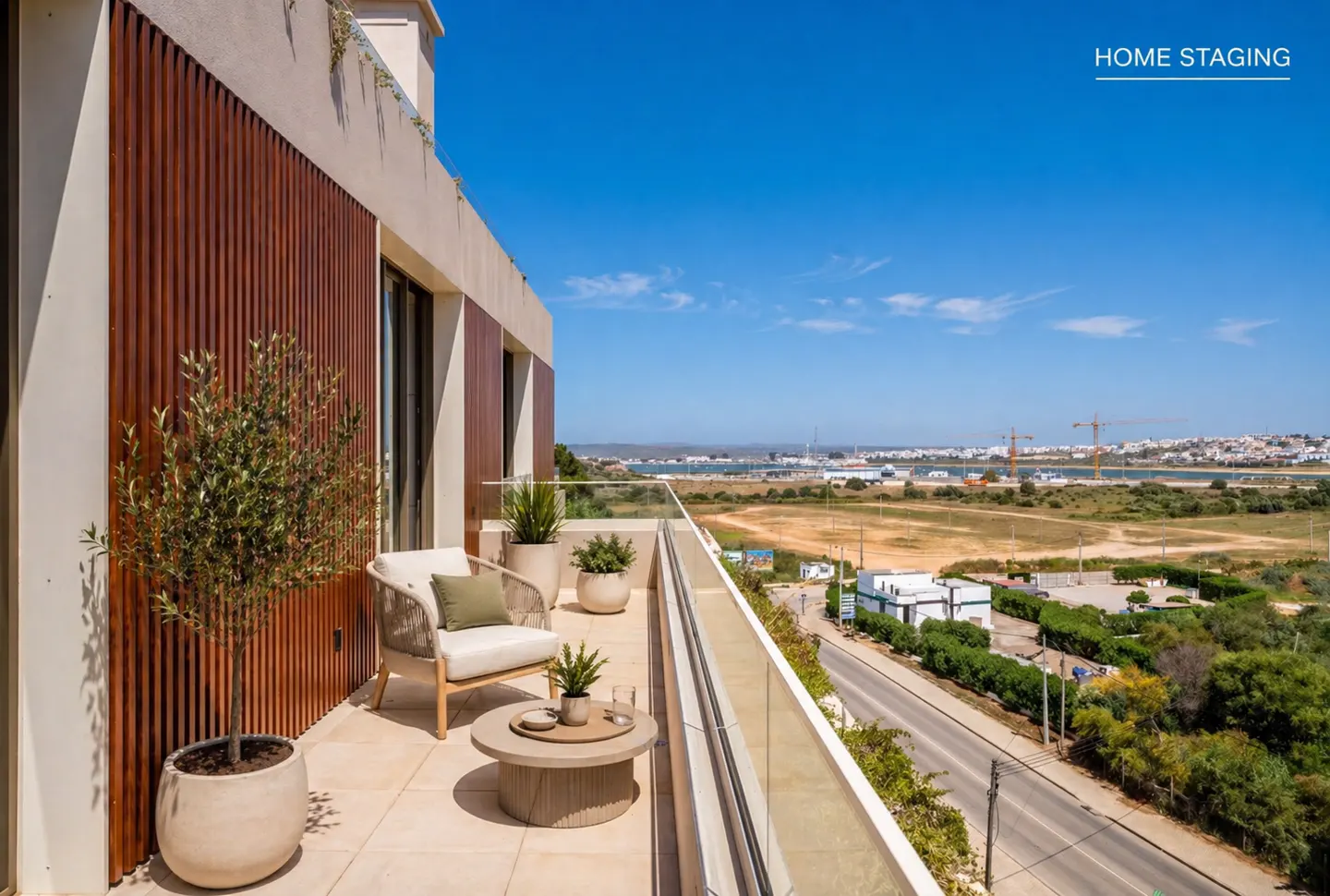 Balcony with chair, table, and potted plants overlooking a city and harbor under a blue sky.