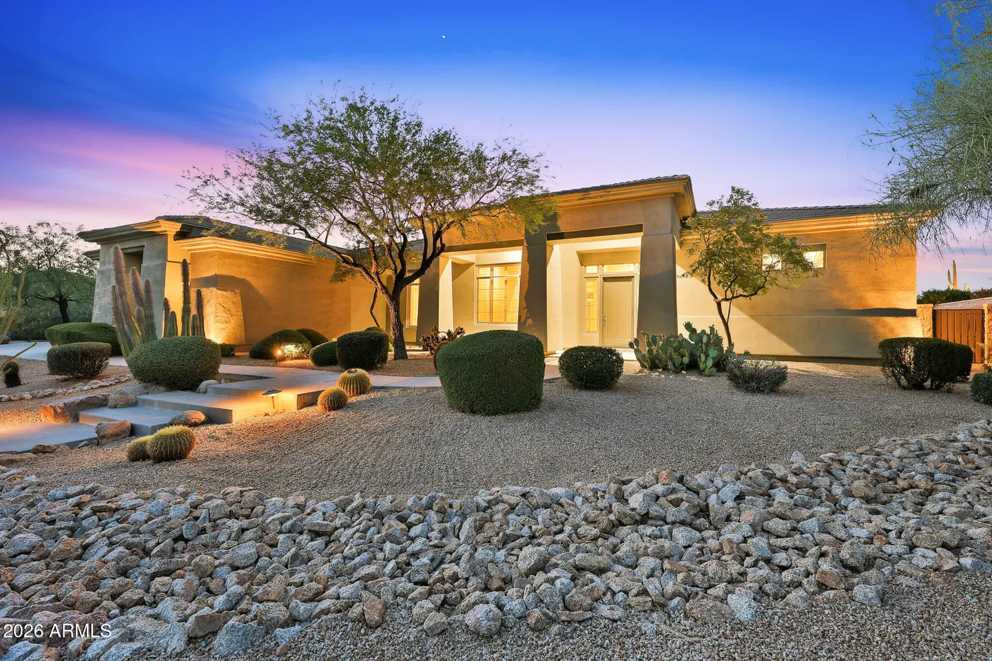 Tan, modern home with desert landscaping at dusk. Cacti, bushes, and trees surround the house. The sky is blue and pink.