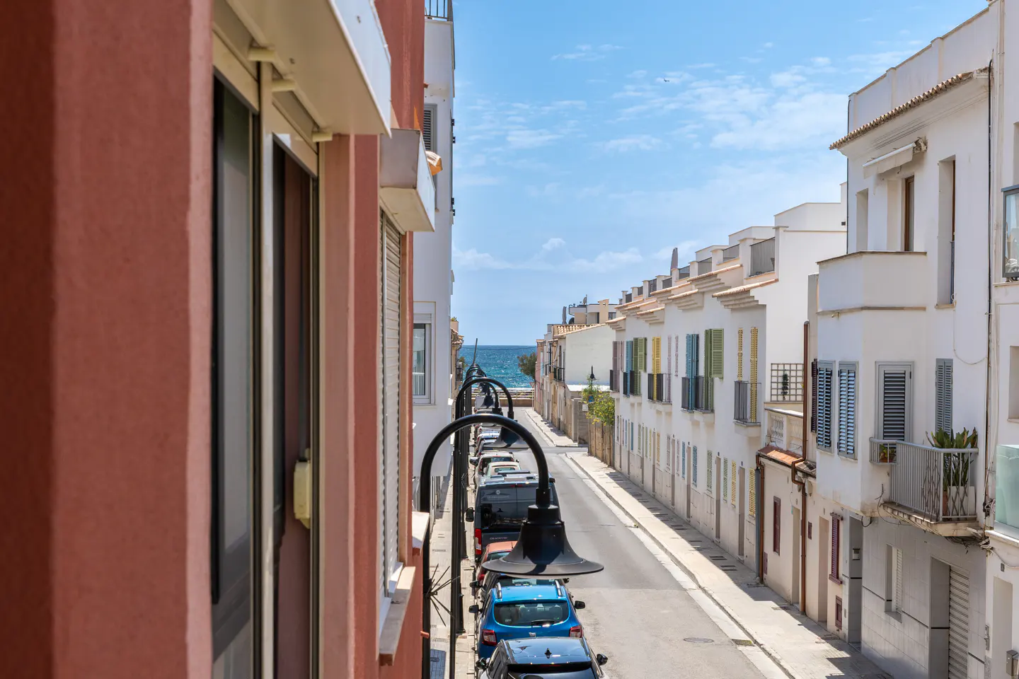 View down a narrow street lined with parked cars and white buildings towards the ocean.