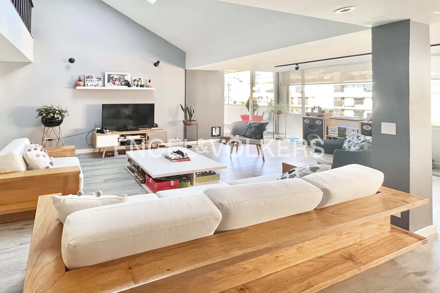 Modern living room with light wood sofa, white coffee table, TV console, and large windows leading to a balcony.