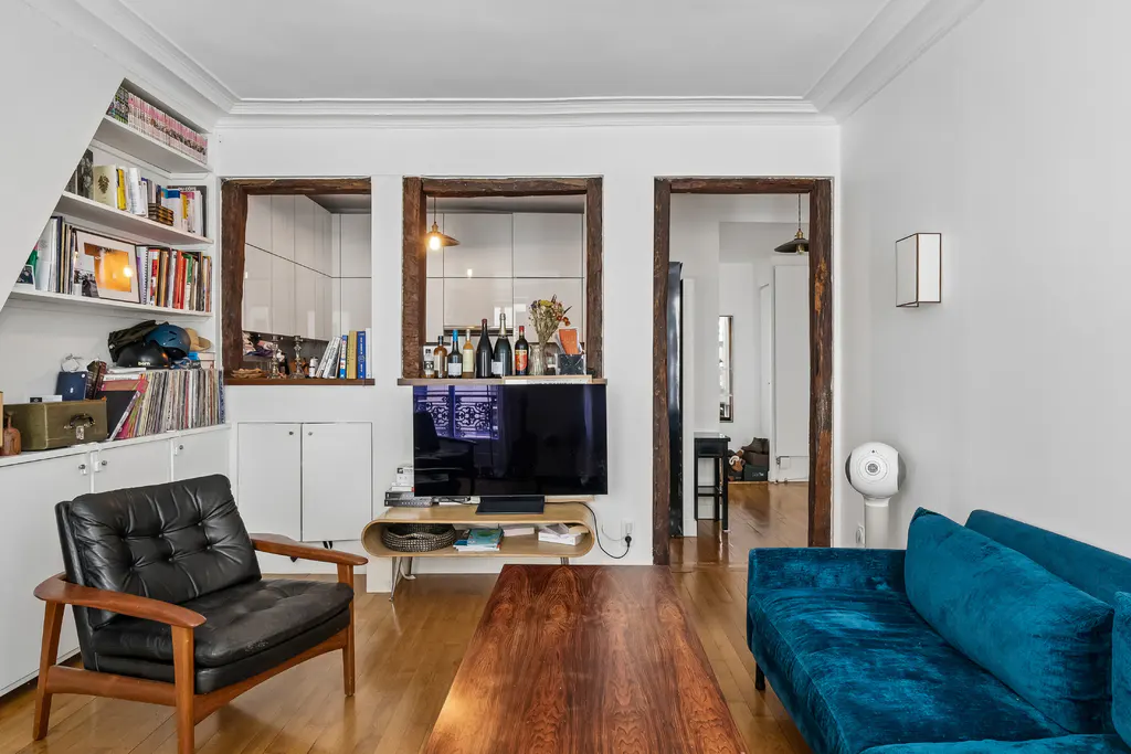 A living room with a blue velvet sofa, a black leather chair, and a wooden coffee table on a wood floor. There is a TV and a bookshelf in the background.