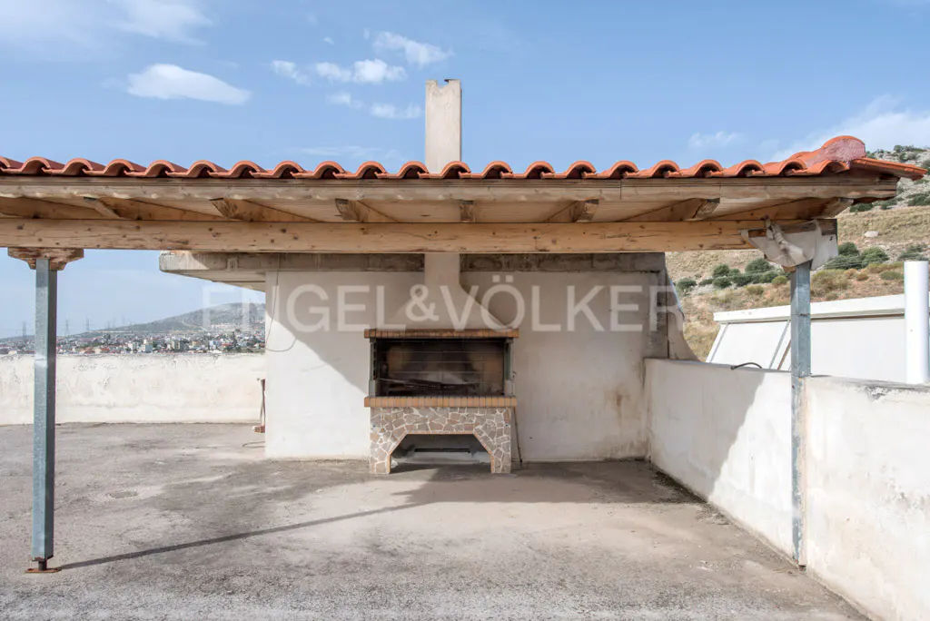 Outdoor rooftop terrace with a covered stone barbecue area, wooden beams, and a view of the distant town and hills.