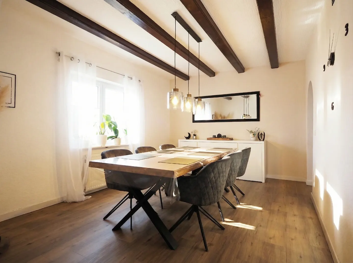 A dining room with a wood table, gray chairs, and dark wood beams on the ceiling. A white sideboard and mirror are in the background.