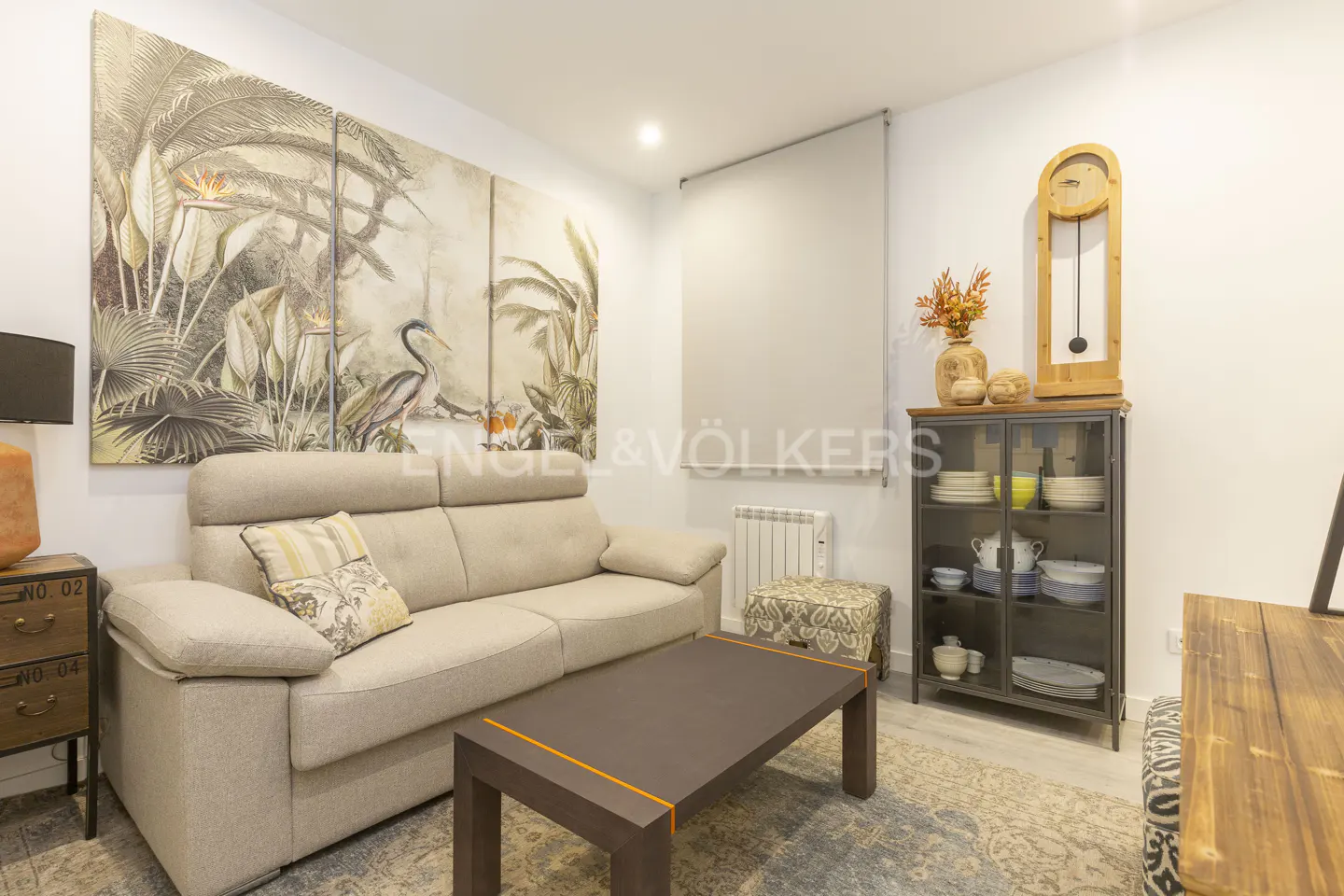 Living room with beige sofa, brown coffee table, and tropical artwork. A glass cabinet and wooden clock are against the white wall.