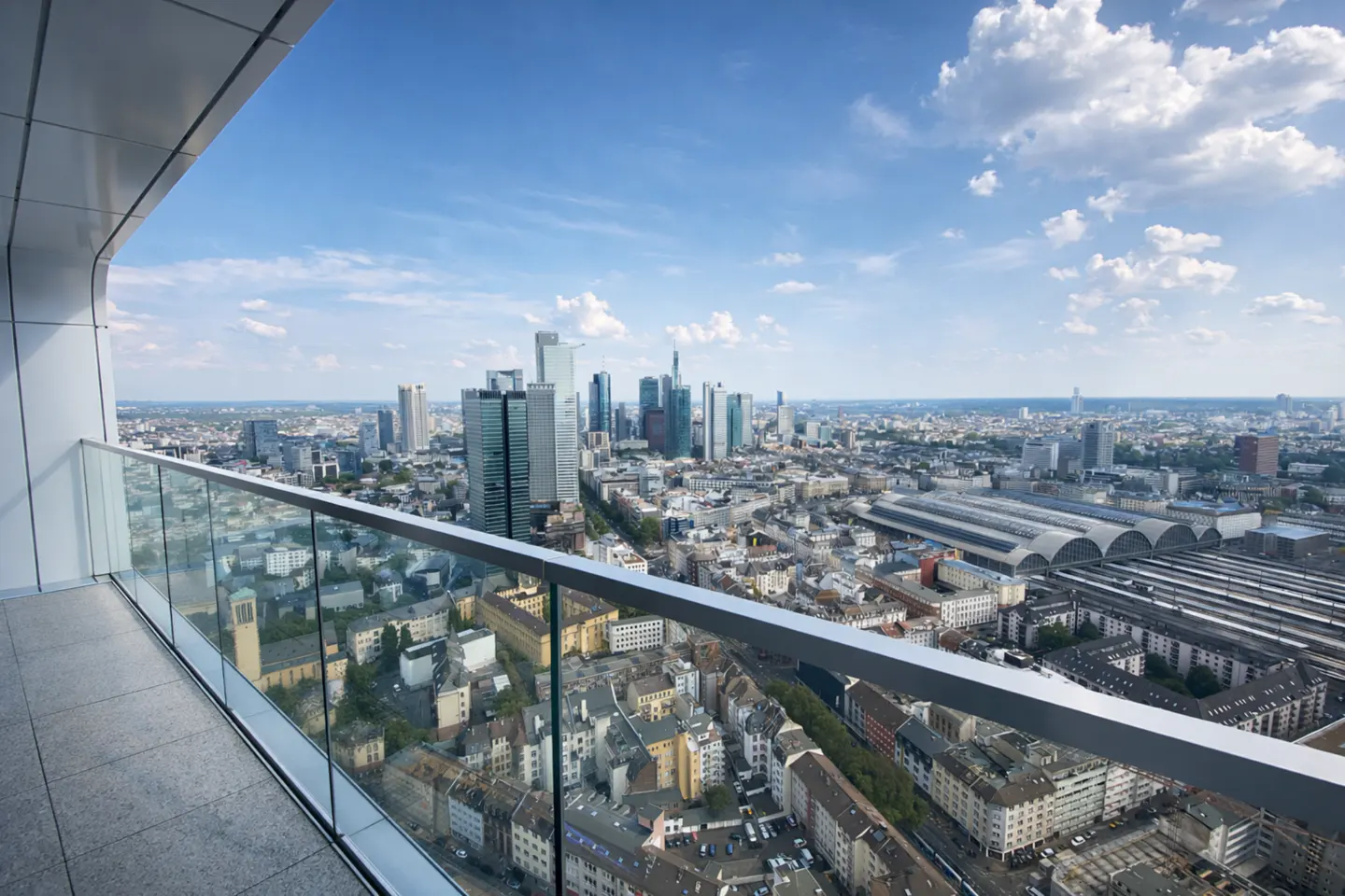 High-rise balcony view overlooking the Frankfurt city skyline and train station under a bright blue sky with scattered clouds.
