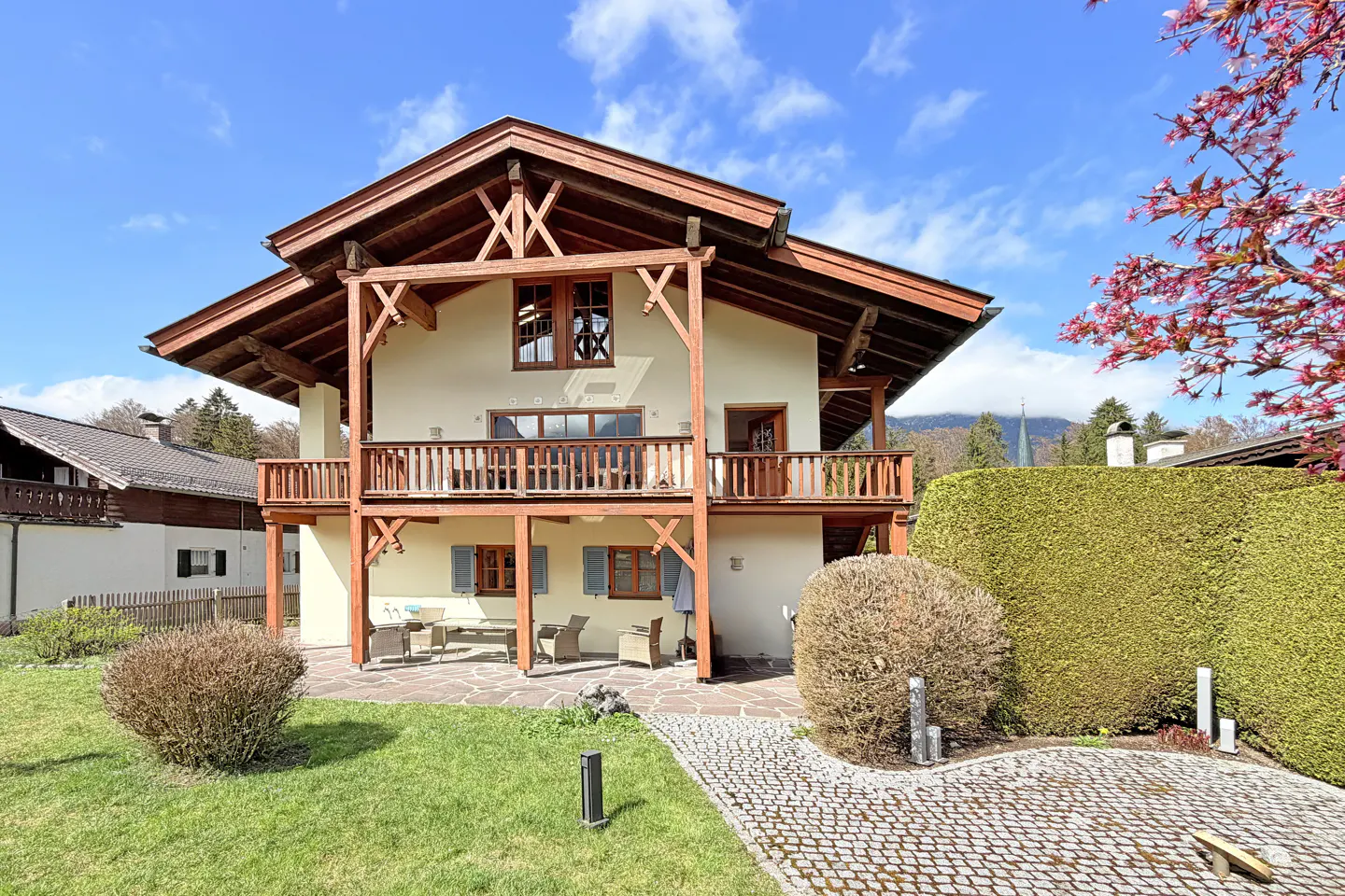 Two-story house with a wooden balcony, brown roof, and light yellow walls. A stone path leads to the house, surrounded by green grass and bushes. Blue sky.