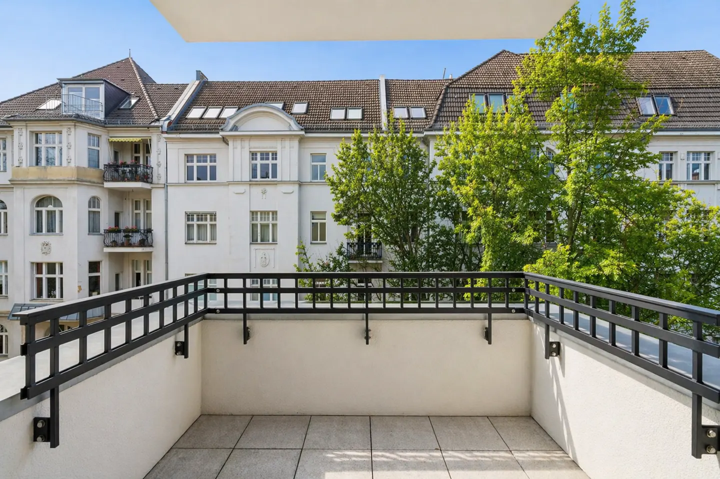 A balcony with a black metal railing overlooks a white building with trees.