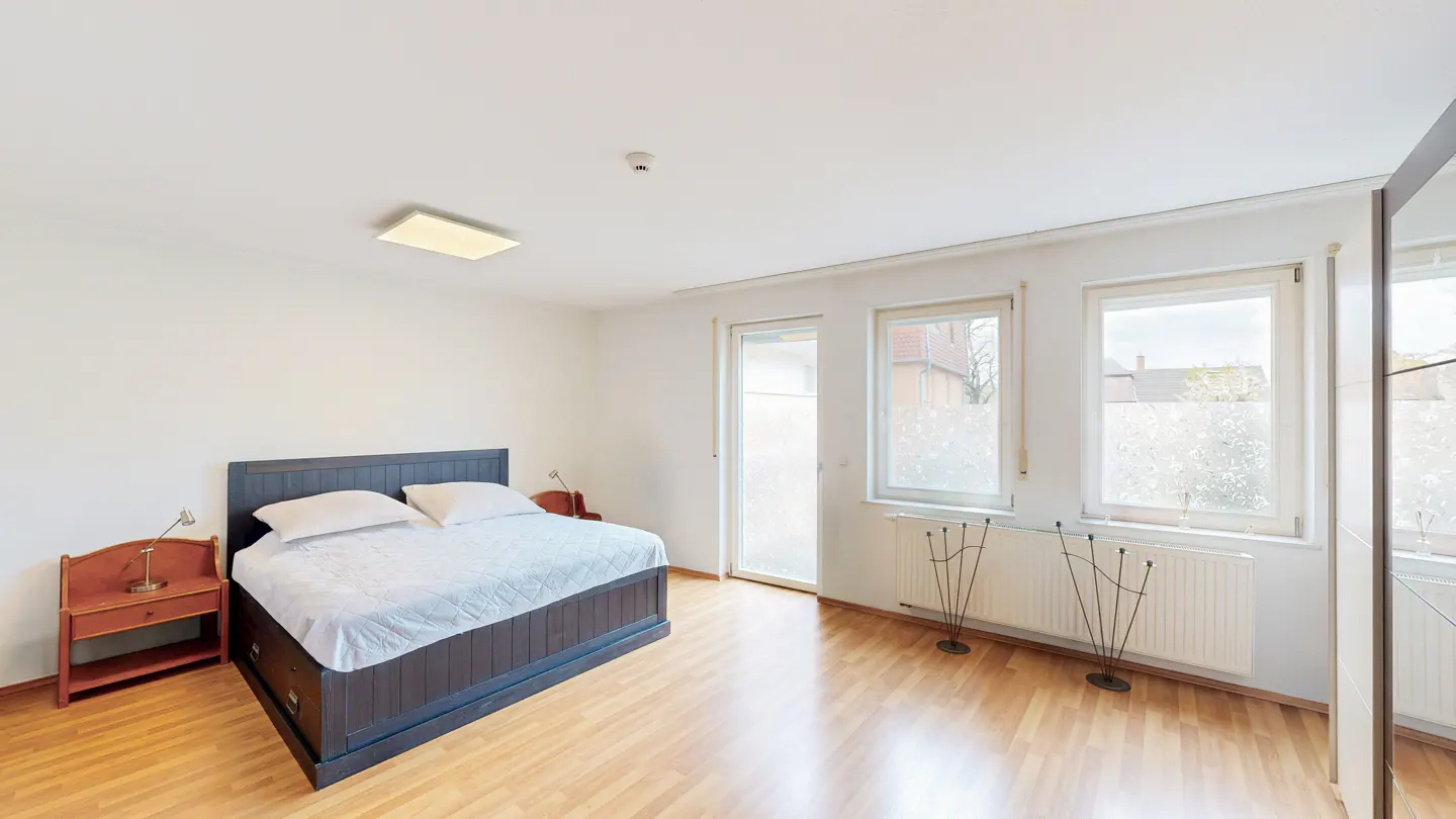 Bright bedroom with a dark wood bed, white linens, and wood nightstands. Windows with frosted glass and wood floors.