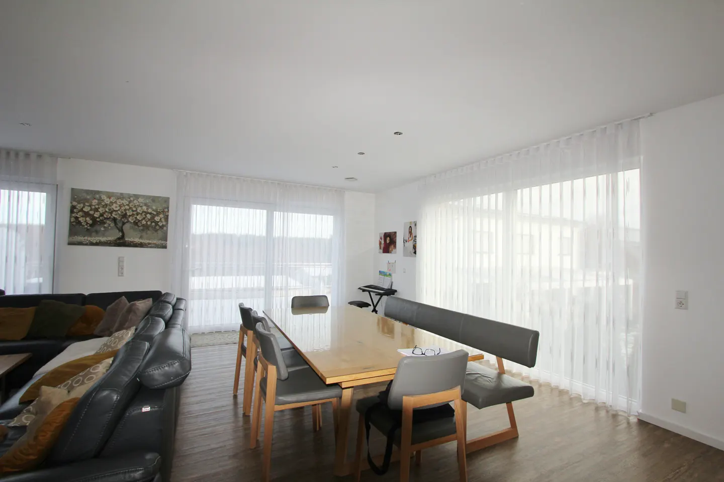 Bright, open-plan living and dining area with a large wooden table and gray chairs. A black leather sofa sits to the left, and sheer white curtains cover the windows.