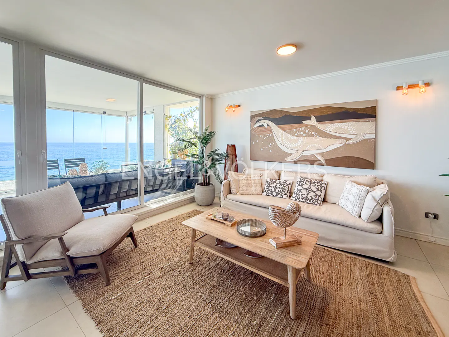 Living room with beige sofa, chair, and jute rug. Ocean view through large windows. Whale art on the wall.