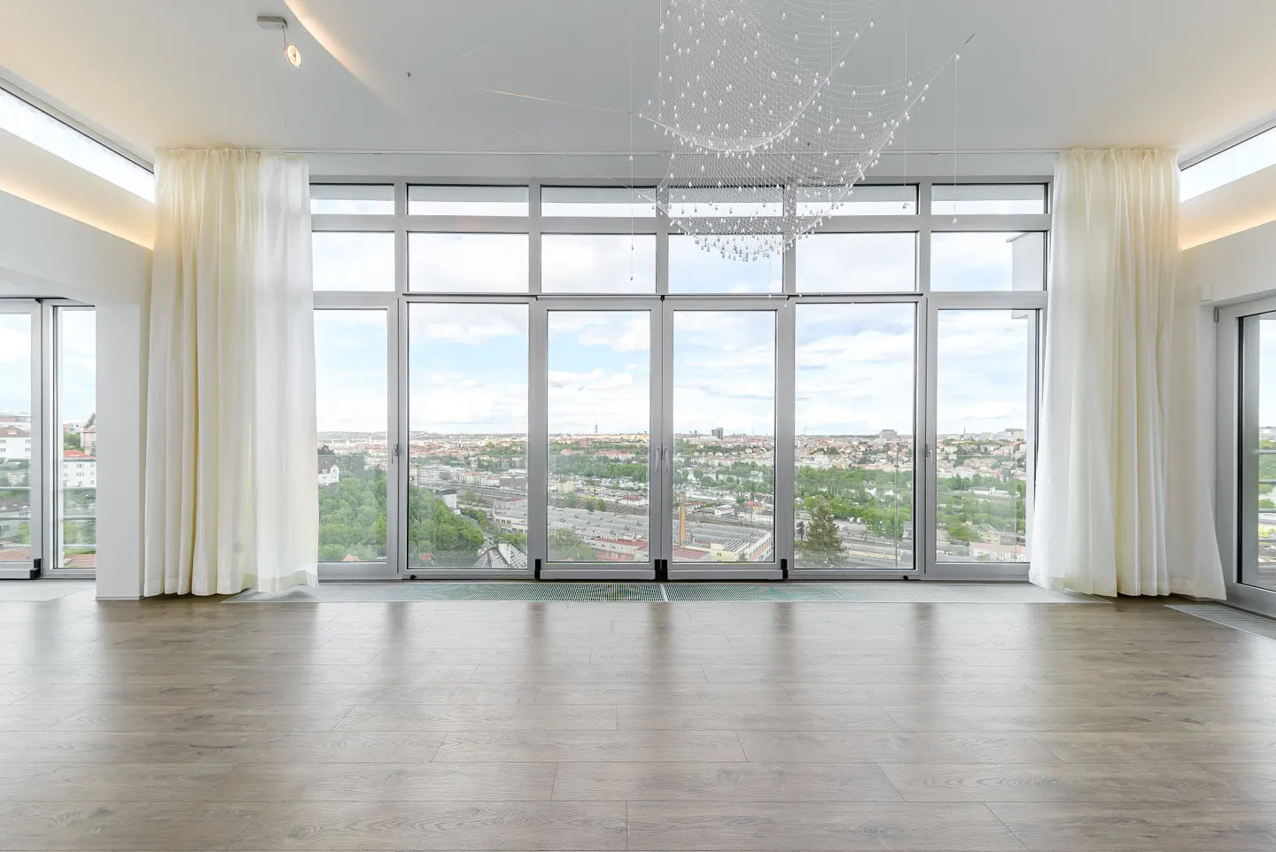 Modern living room with wood floors, white curtains, and a large glass wall offering a panoramic city view.