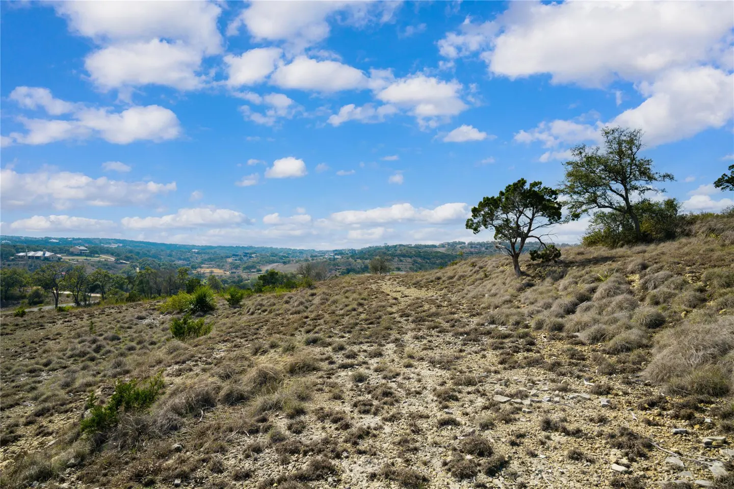 Hilly landscape with sparse vegetation under a blue sky with scattered clouds. Trees dot the hillside, and houses are visible in the distance.