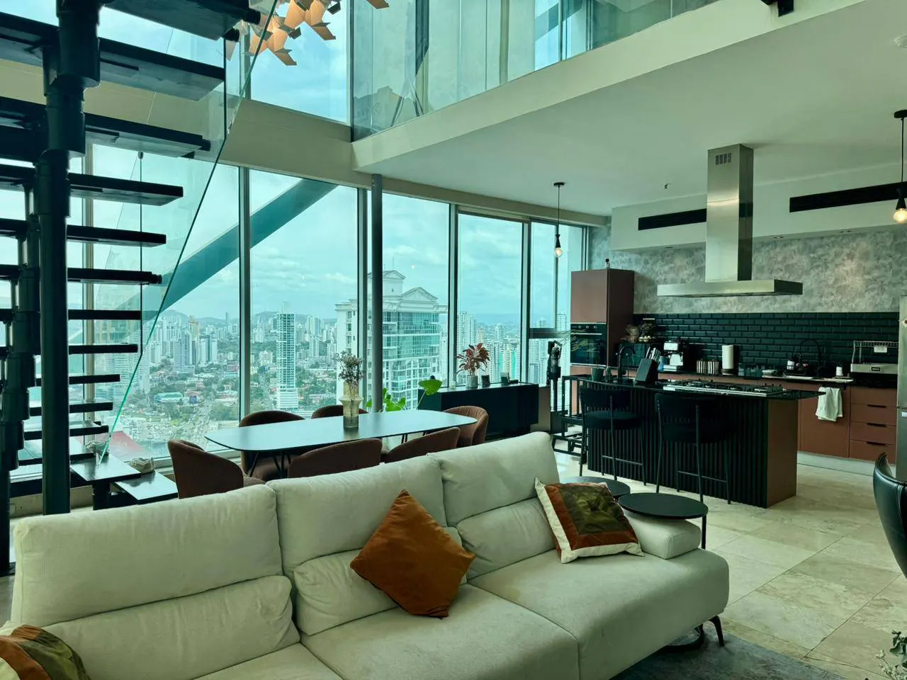 Bright, modern apartment interior with floor-to-ceiling windows overlooking a city. A white sofa, dining table, and kitchen are visible.