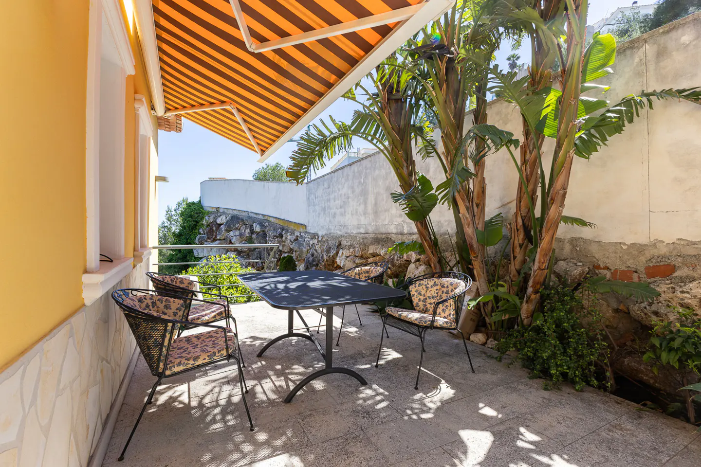 Outdoor patio with a black table and four chairs with floral cushions, under a striped orange awning, next to a yellow building and palm trees.