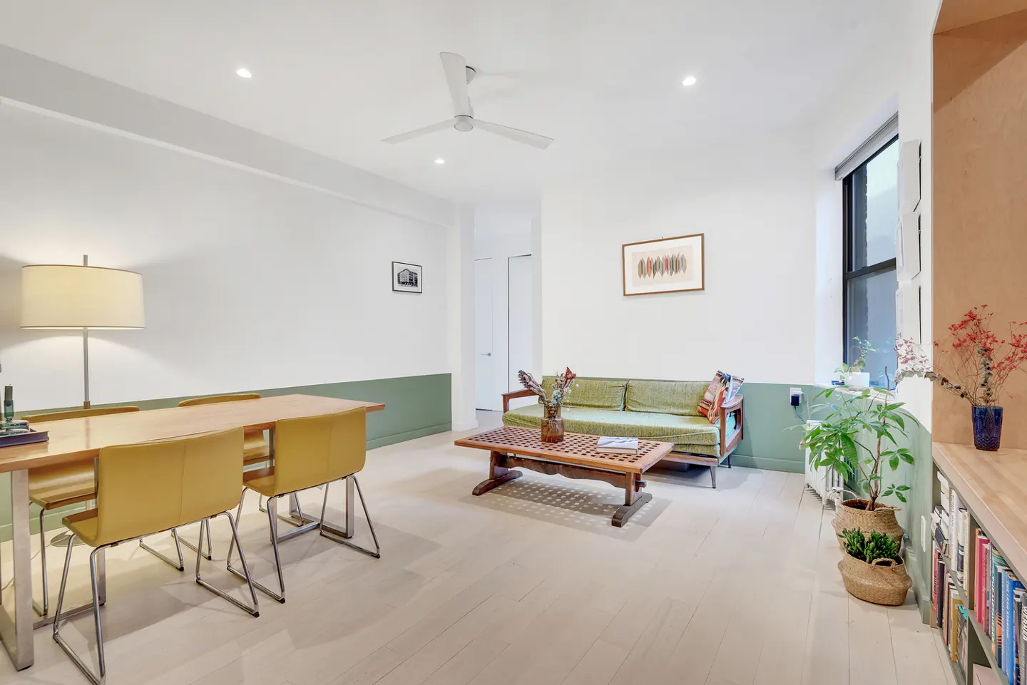 Bright living room with white walls, green trim, and light wood floors. A green sofa, wood table, and yellow chairs furnish the space.