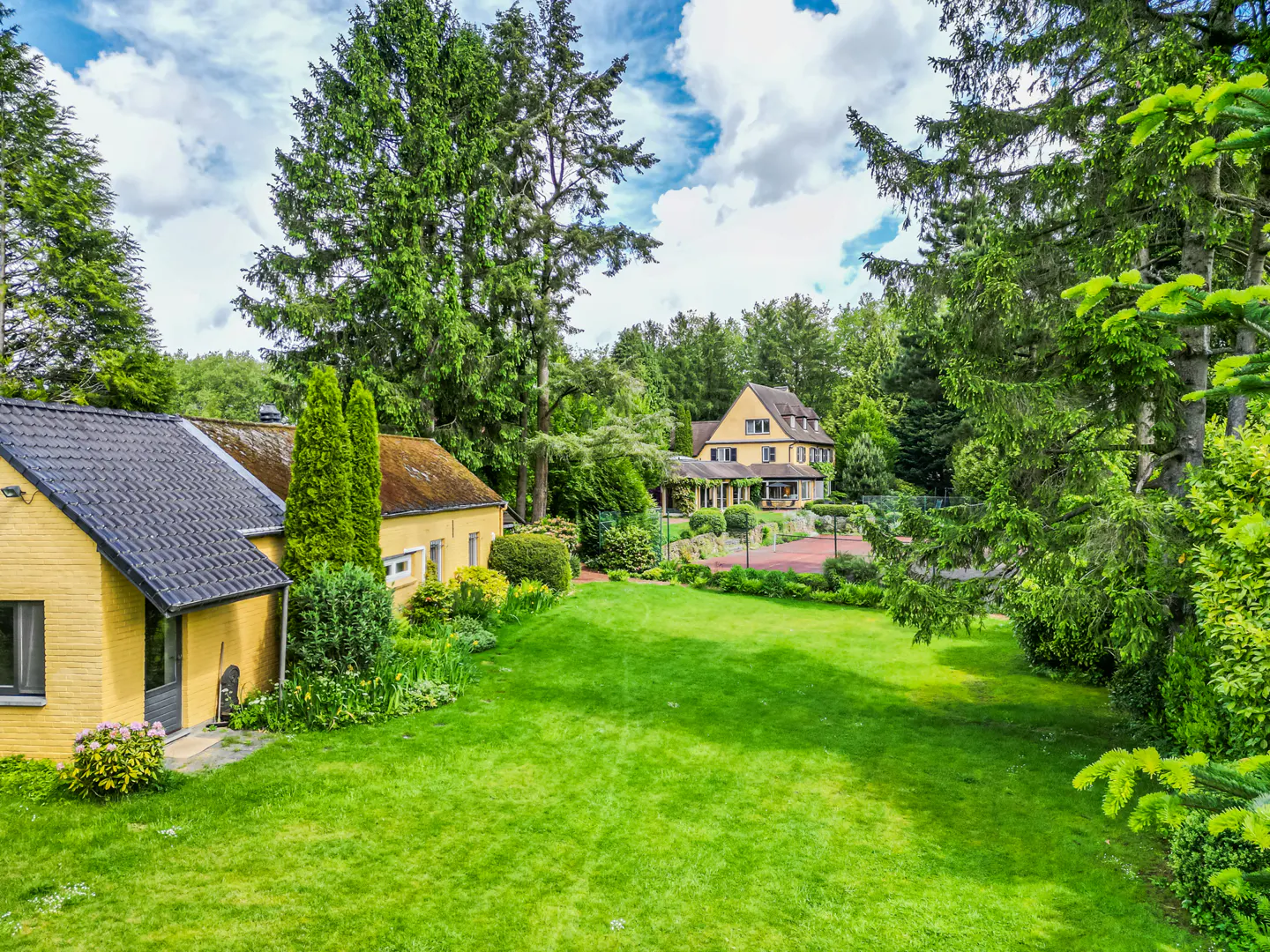 A yellow house with a black roof sits on a green lawn surrounded by trees.
