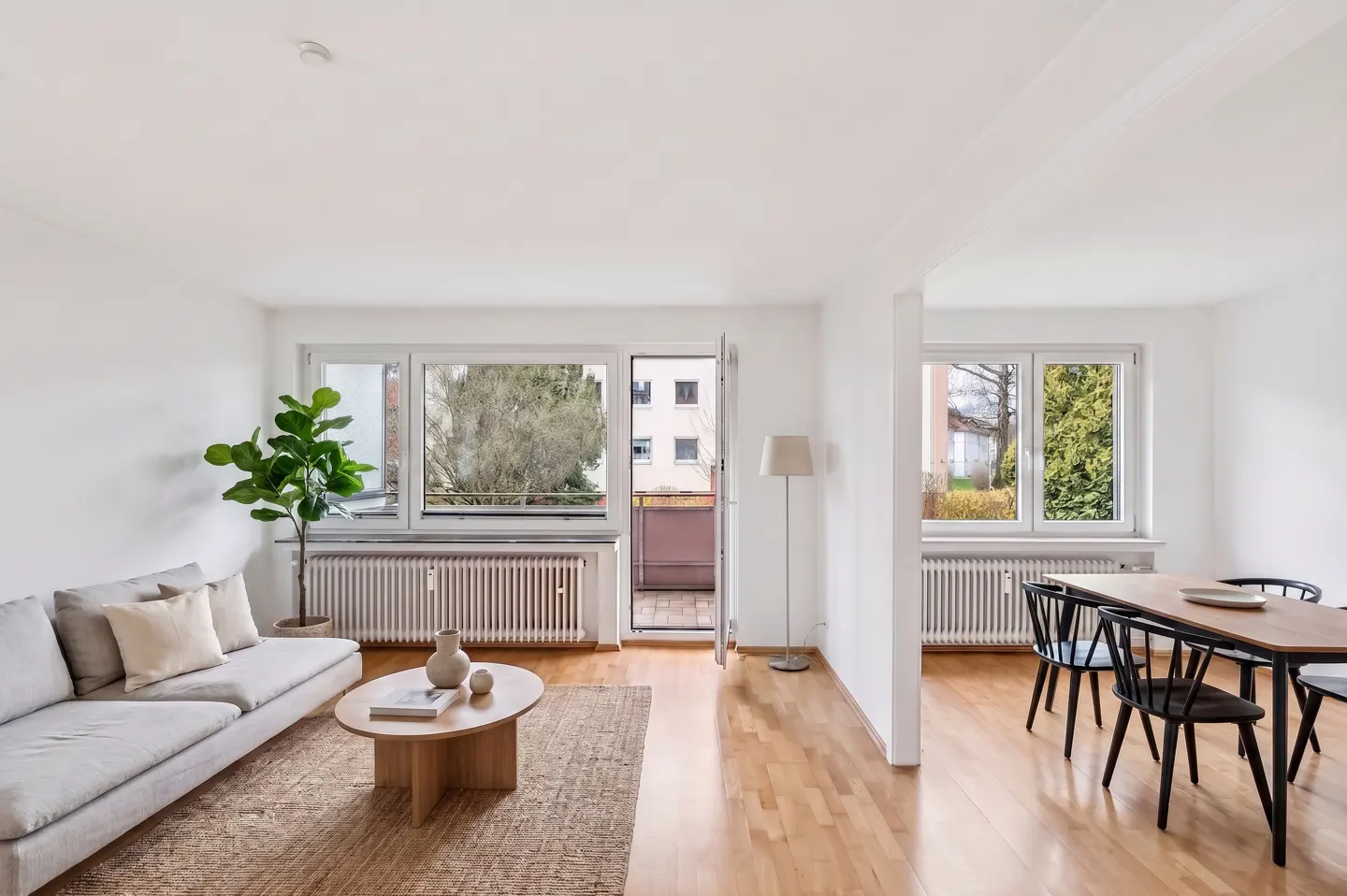 Bright, open-concept living and dining area with hardwood floors, white walls, and large windows. A gray sofa, round coffee table, and dining set furnish the space.