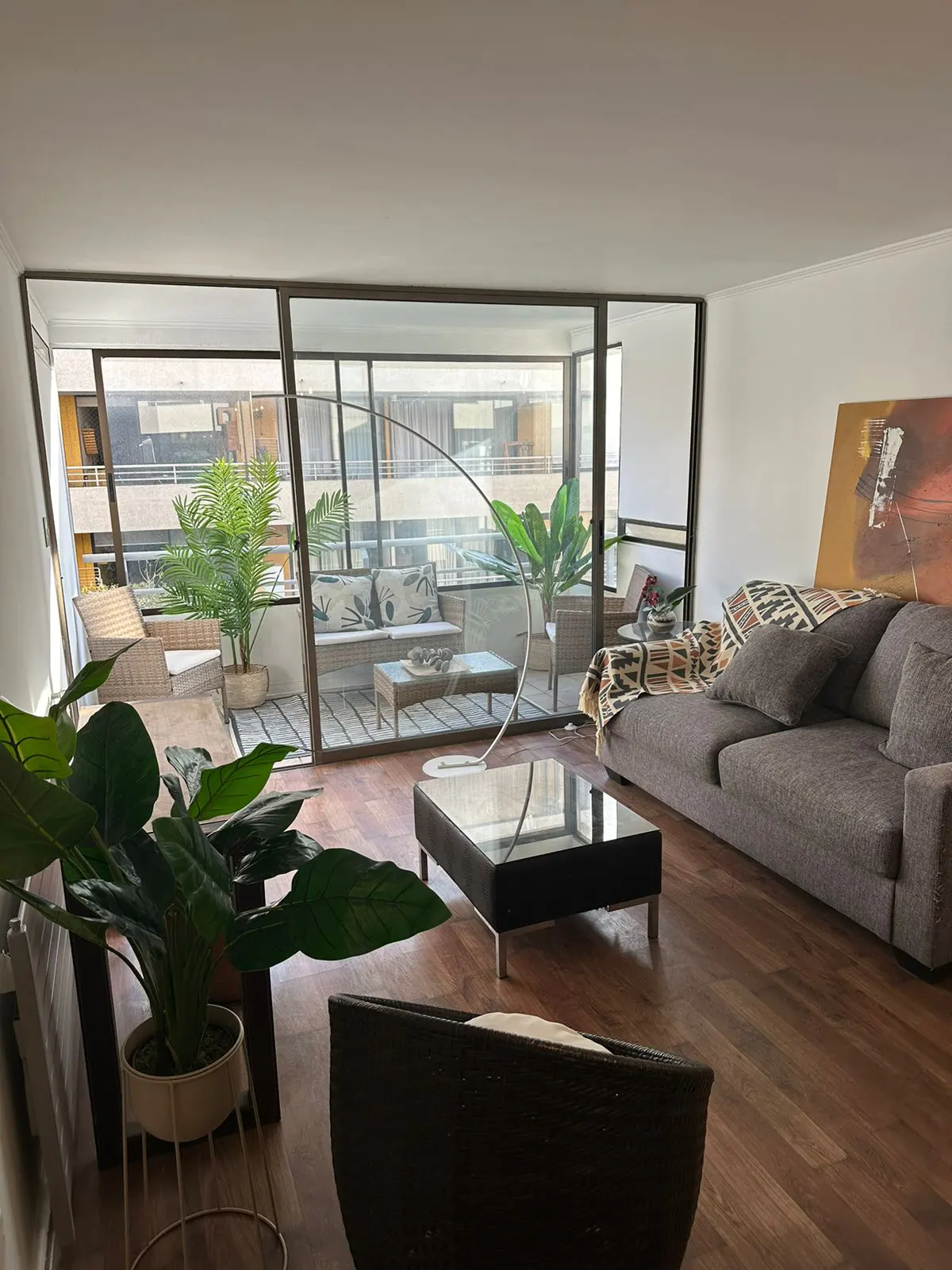 Living room with wood floors, gray sofa with throw, glass coffee table, and balcony with seating visible through large windows.