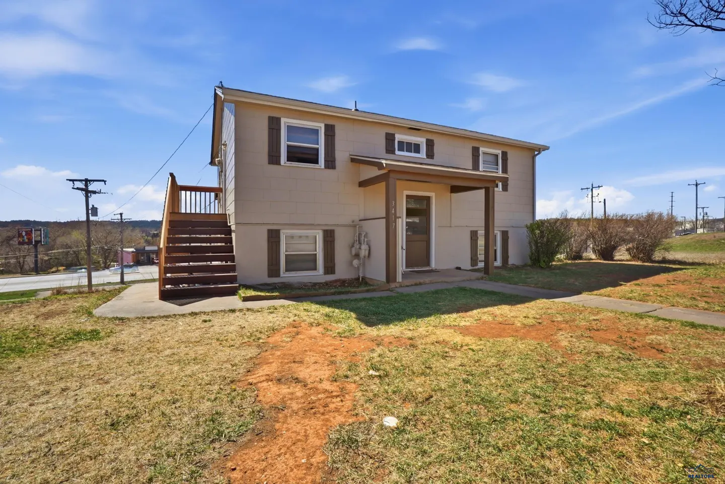 Two-story beige house with brown shutters, a front porch, and exterior wooden stairs under a bright blue sky.