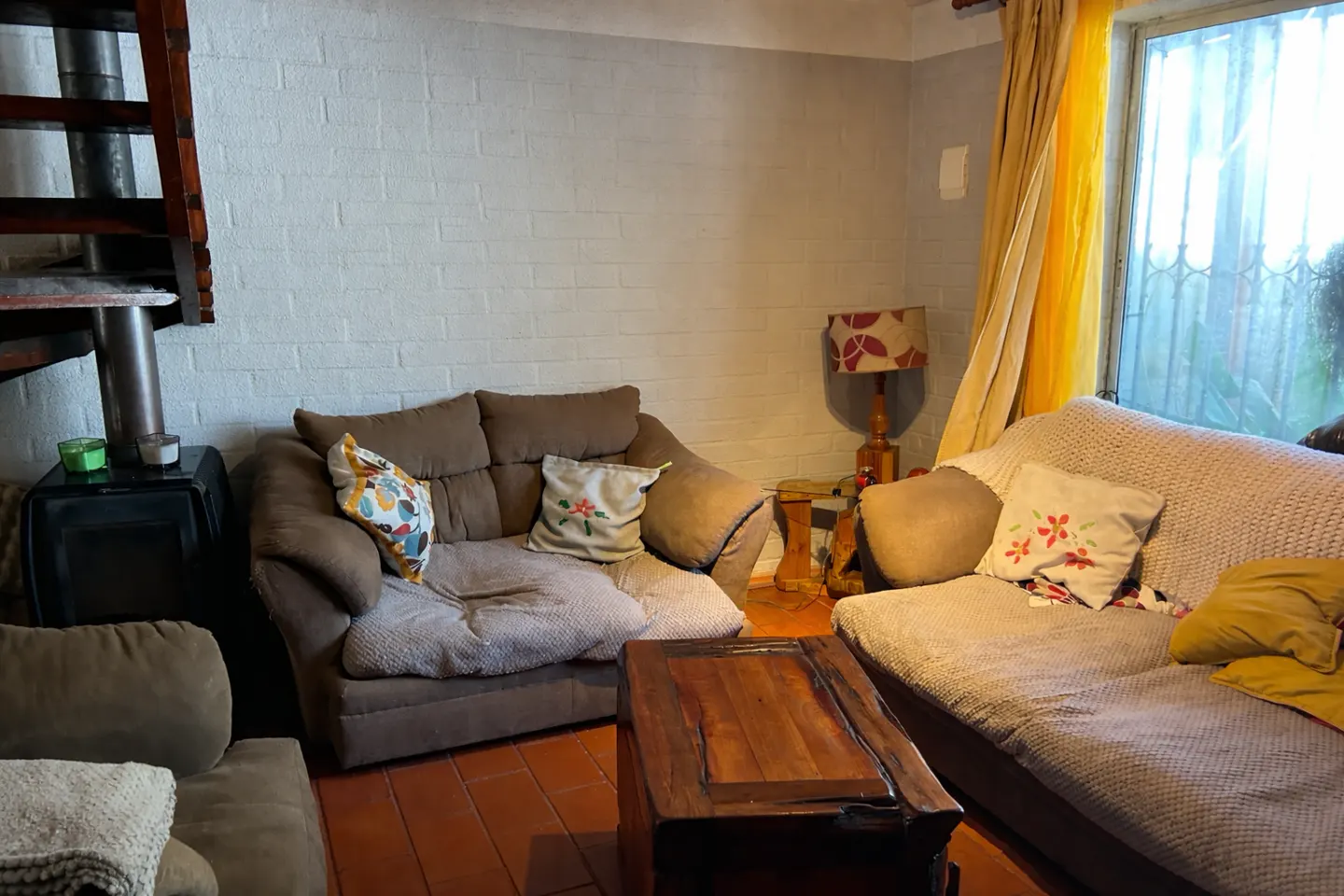 Cozy living room with brown sofas, wooden coffee table, and a wood-burning stove near a spiral staircase. Yellow curtains frame a bright window.