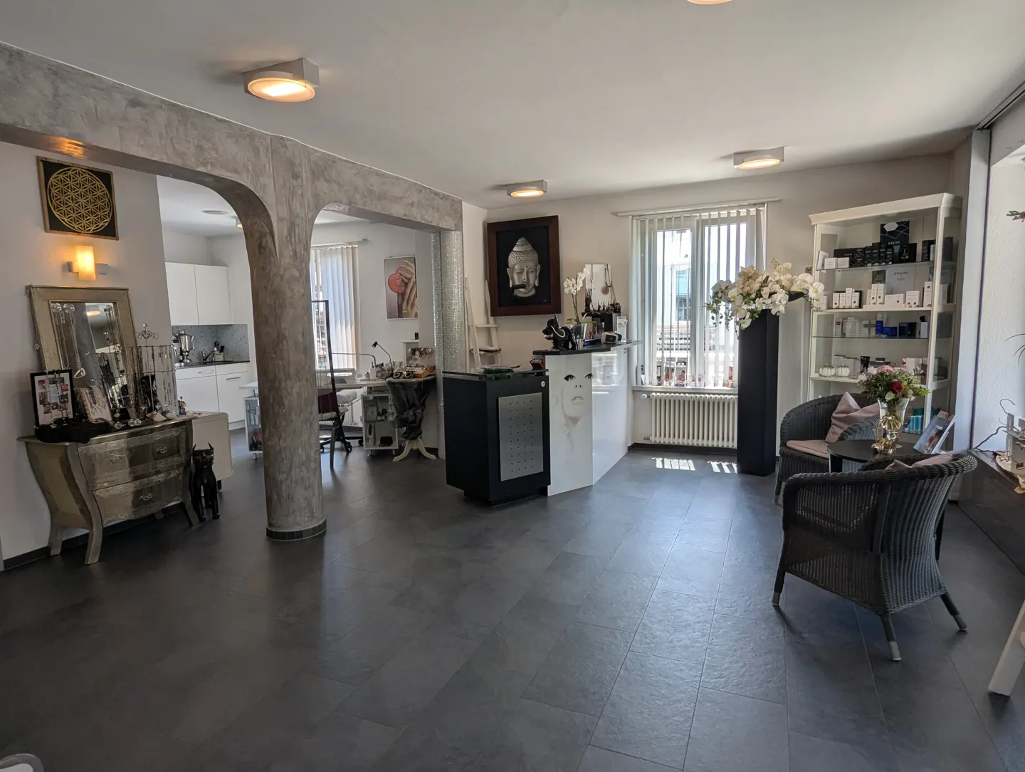 Interior view of a beauty salon with gray floors, white walls, and a reception desk. There are chairs, shelves with products, and decorative items.