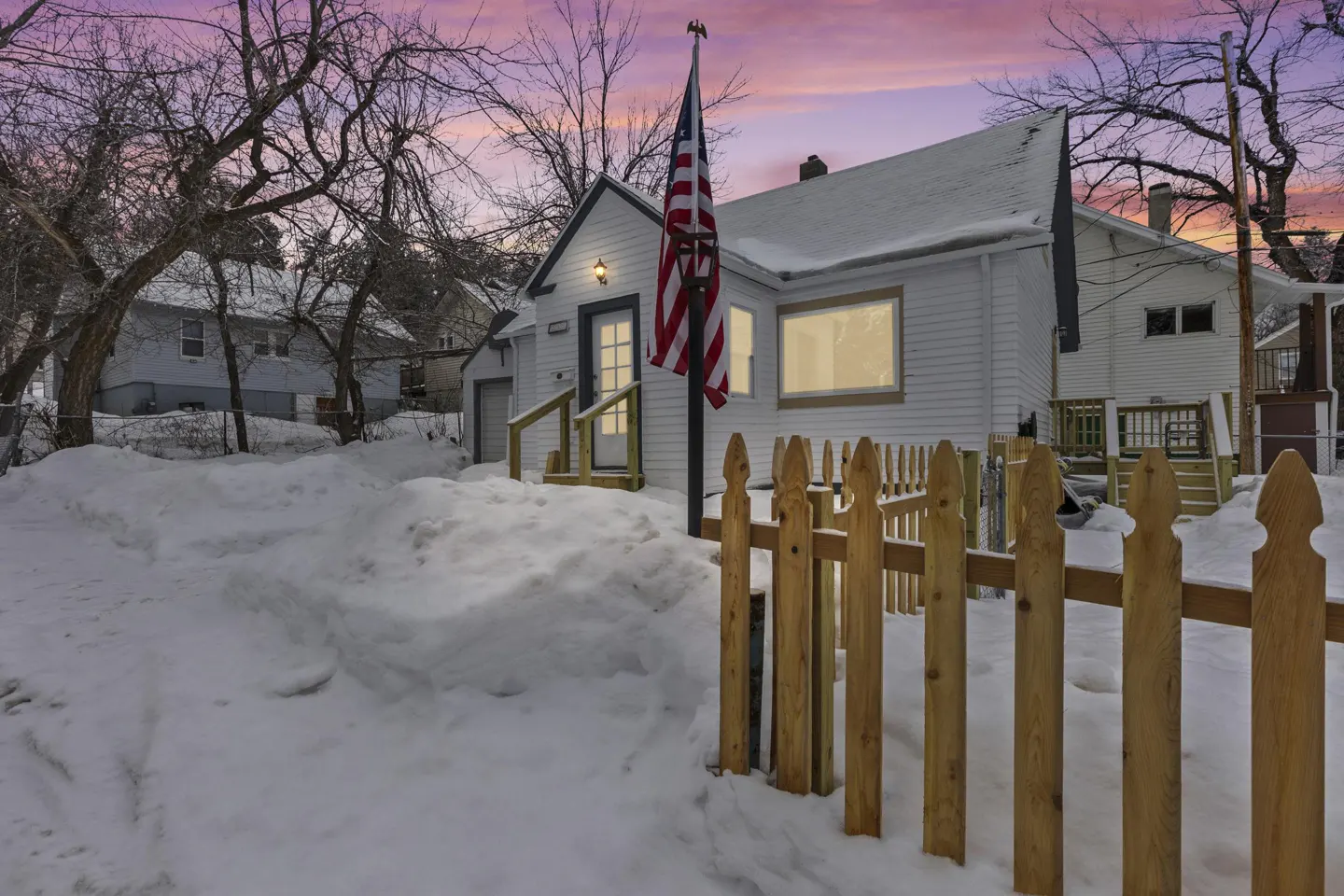 Exterior of a white house with snow, a wooden fence, and an American flag at dusk.