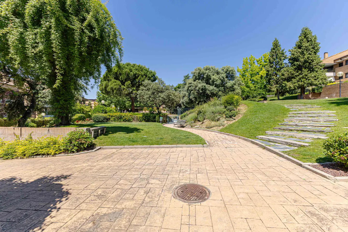A sunny park view with stone paths, green grass, and trees. A stone staircase leads up a grassy hill. A building is visible in the background.