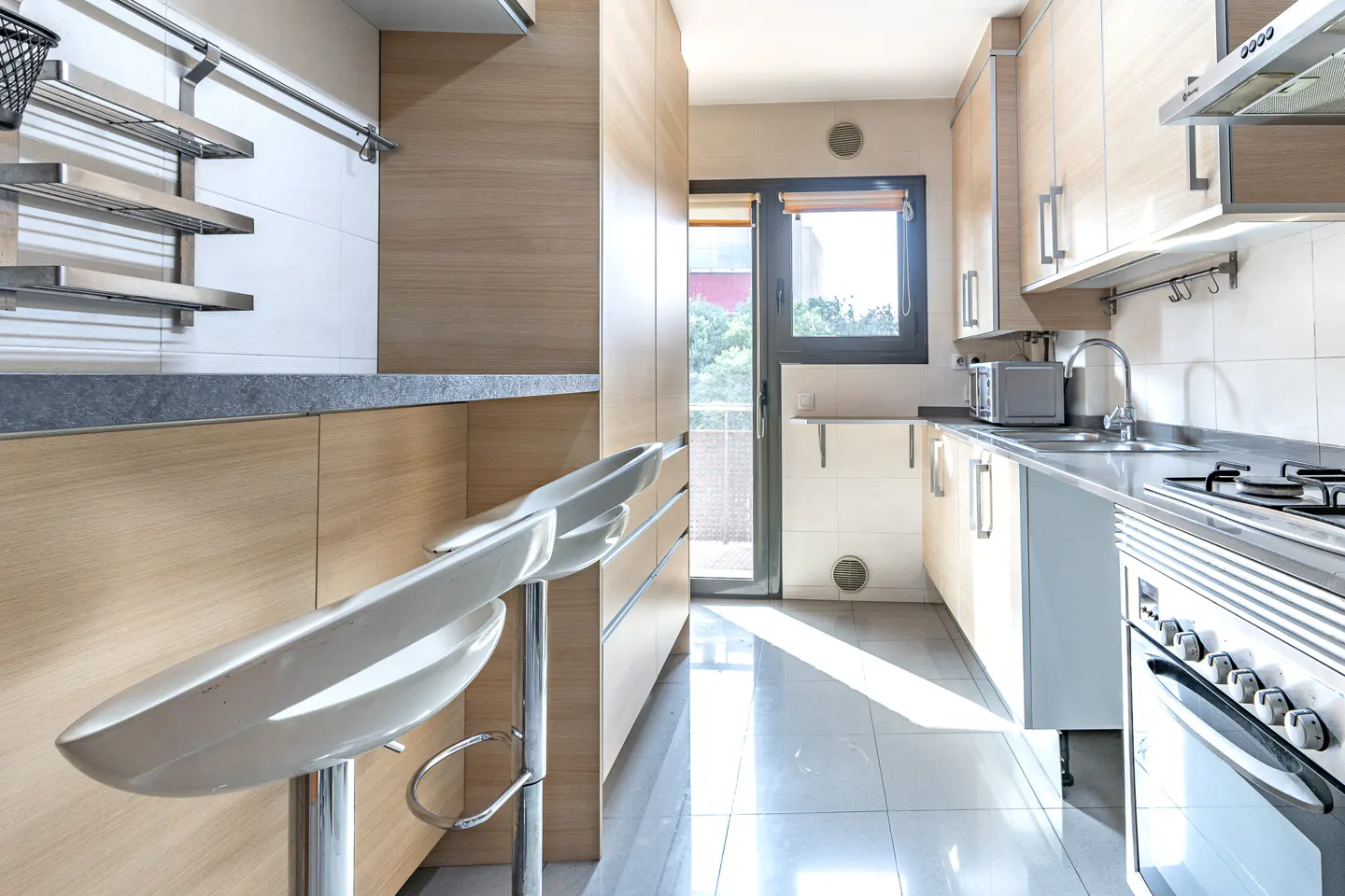 Modern galley kitchen with light wood cabinets, stainless steel appliances, white bar stools, and a glass door to a balcony.