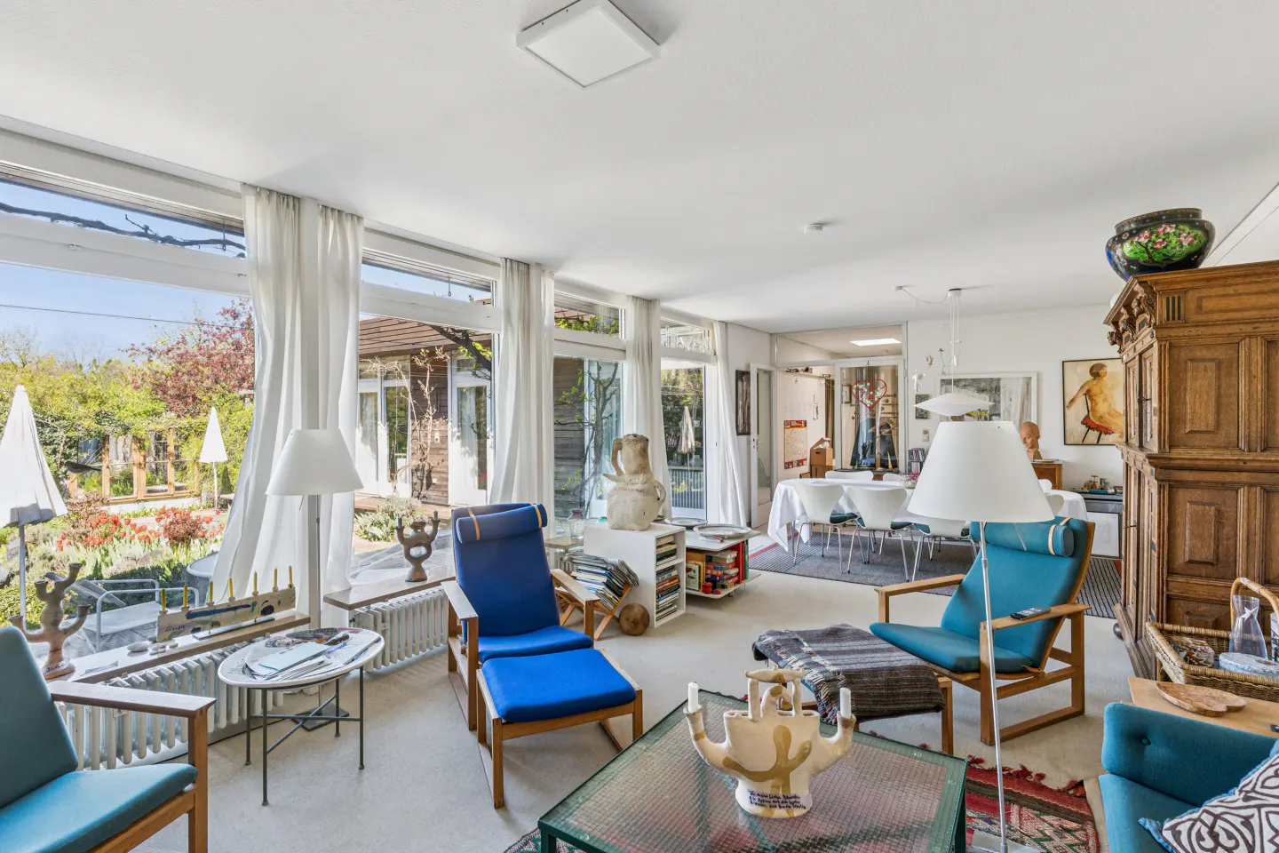 Bright living room with floor-to-ceiling windows, blue armchairs, a coffee table with art, and a large wooden cabinet.