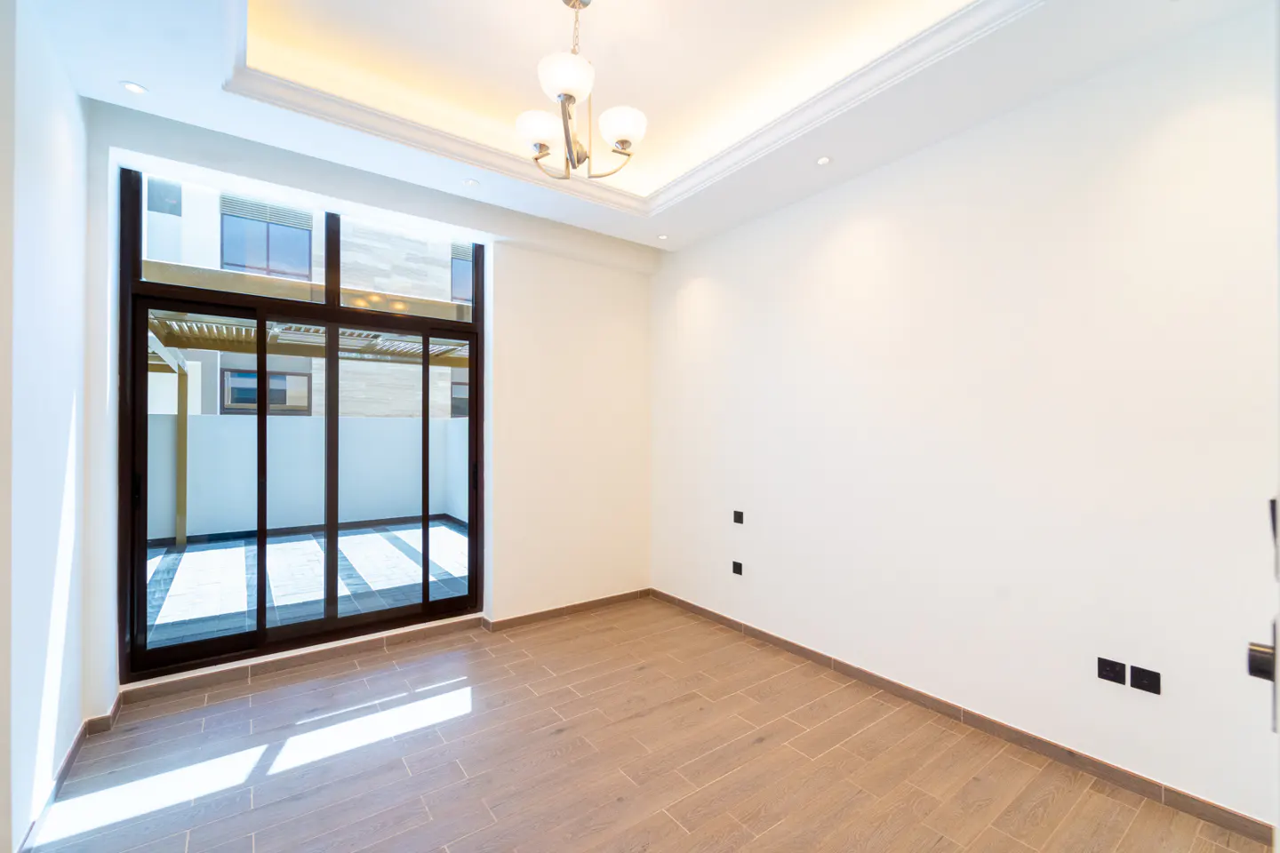 Bright, empty room with wood-look tile floor, white walls, and a black-framed sliding glass door to an outdoor patio. A chandelier hangs from the ceiling.