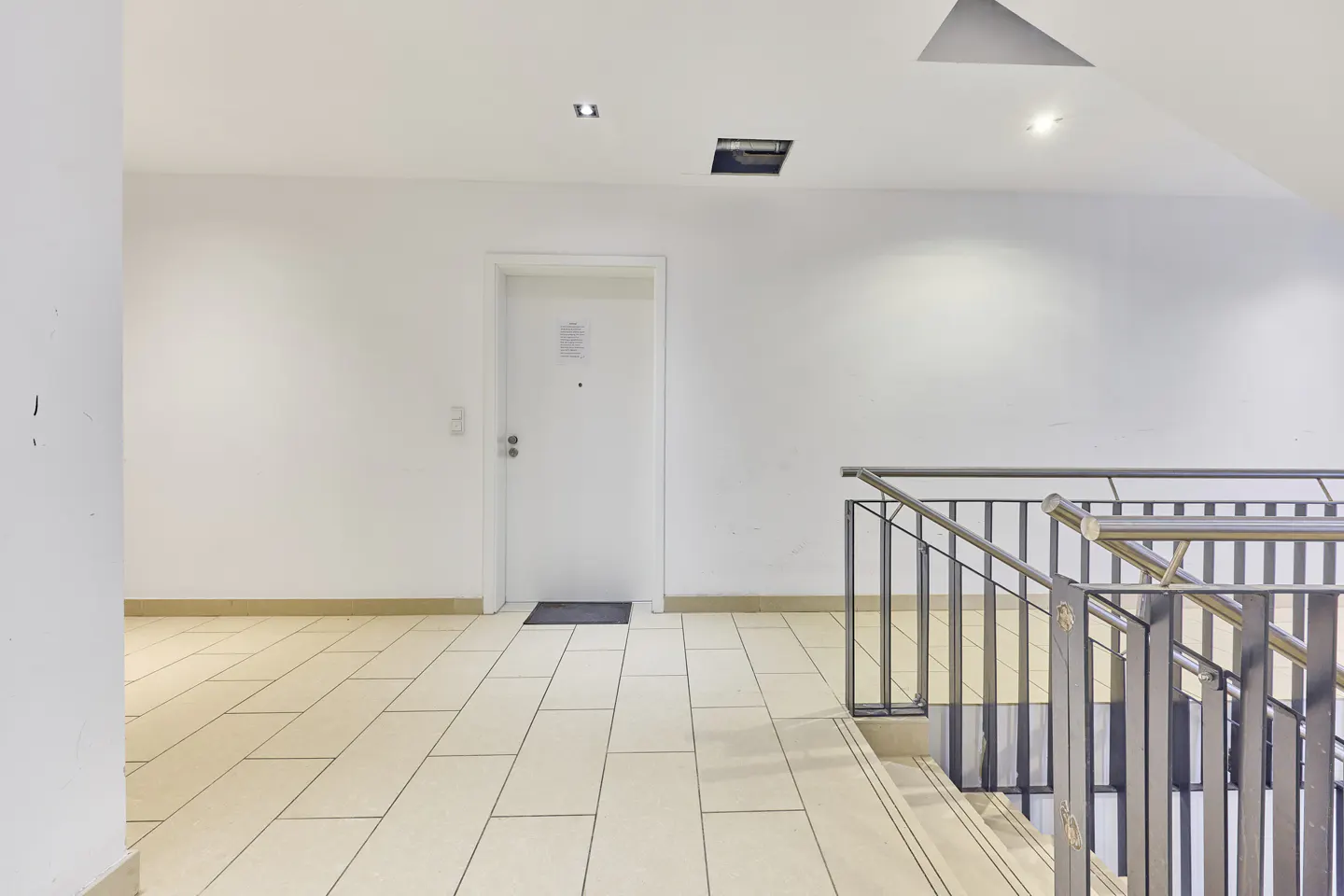 A white door at the end of a tiled hallway next to a metal staircase railing in a bright, minimalist interior.