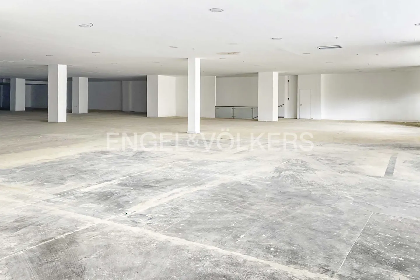 Empty commercial space with white walls, concrete floor, and support columns. Engel & Völkers logo in the center.