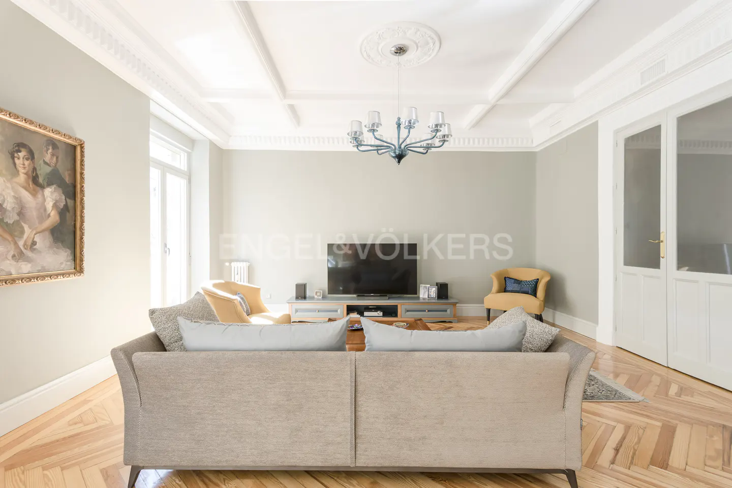 Living room with a beige sofa, TV, and yellow chairs. A blue chandelier hangs from the decorative ceiling. Hardwood floors add warmth.
