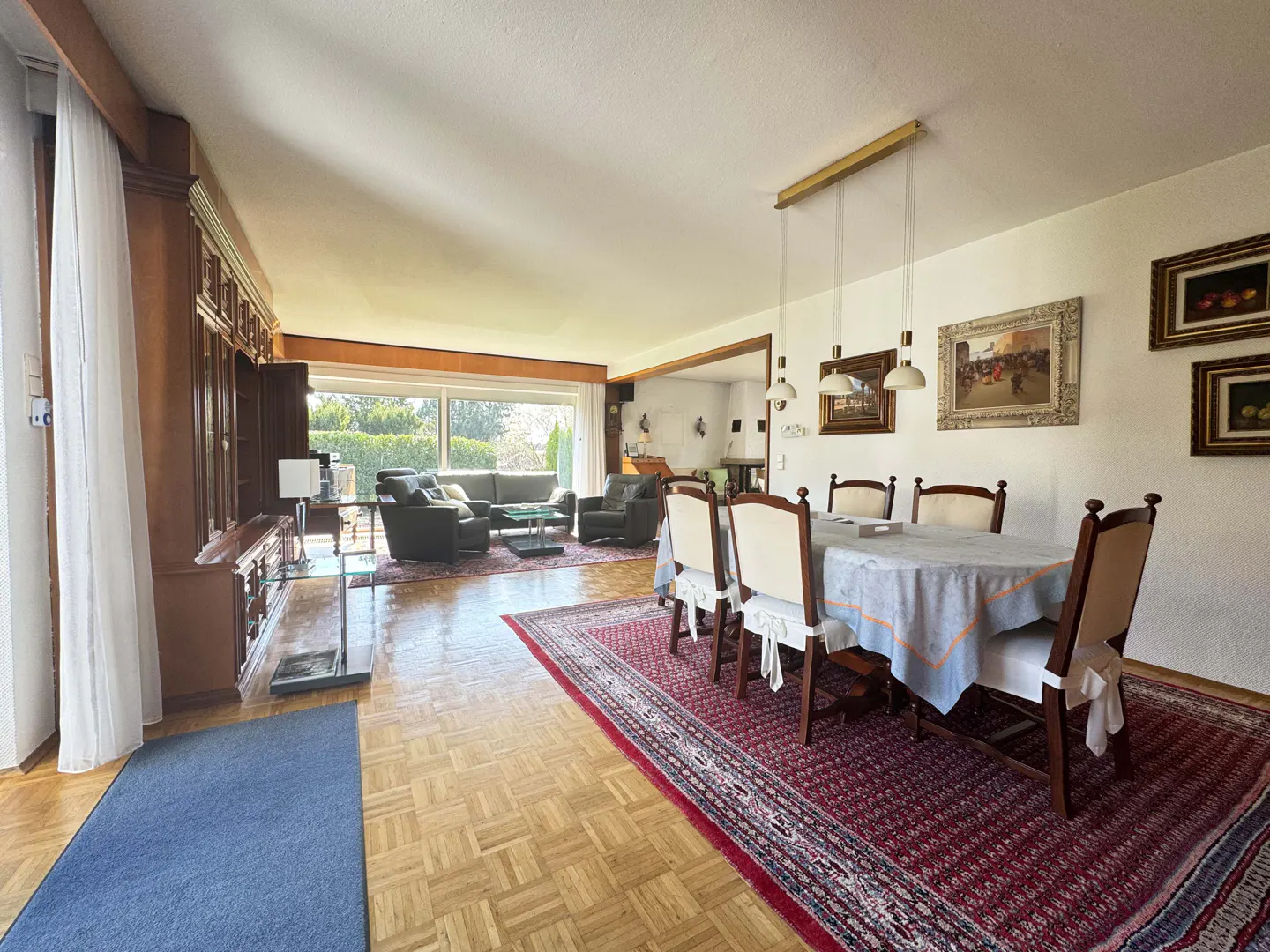 A bright, open-concept living and dining area with hardwood floors and a red patterned rug. A dining table with chairs sits near framed artwork. A living room with sofas is visible in the background.